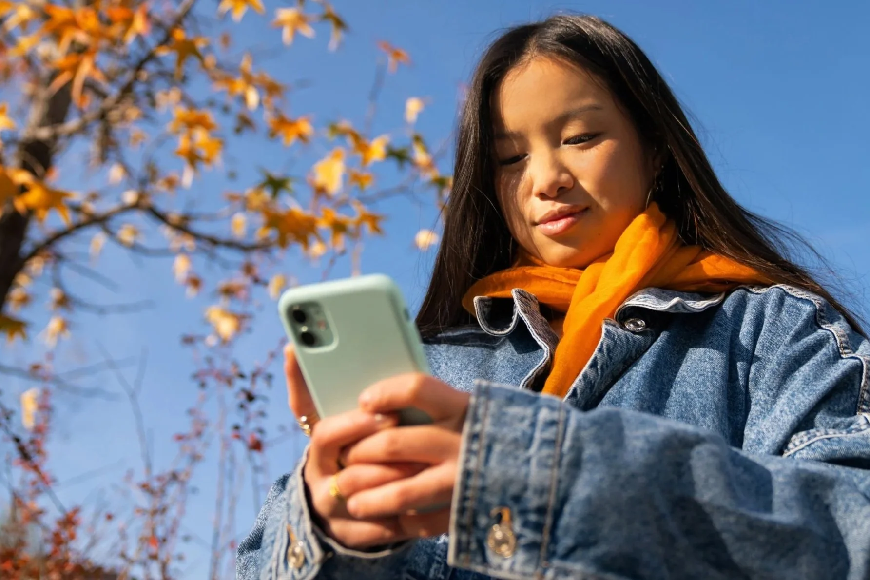 A young woman with long black hair stands outside looking down at her phone. There is a tree with leaves turning orange against a bright blue sky in the background.