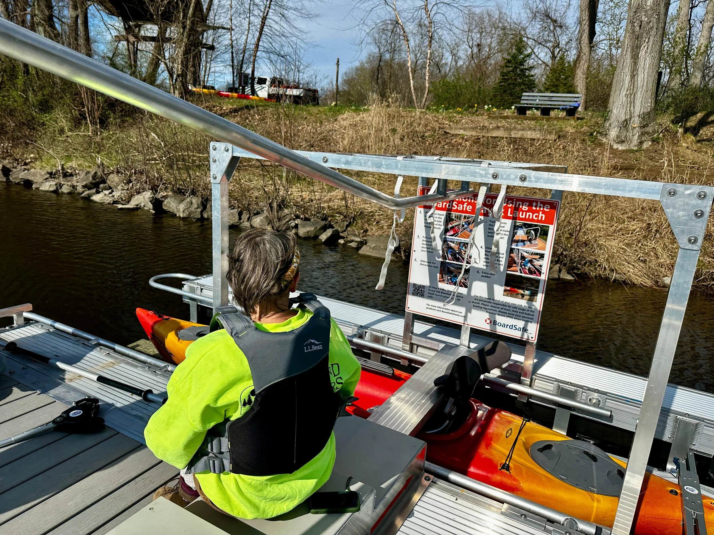A person in a bright green shirt sits on the steps that are part of the gradual entry to the kayak. Above him is a rail to help him ease his way down.