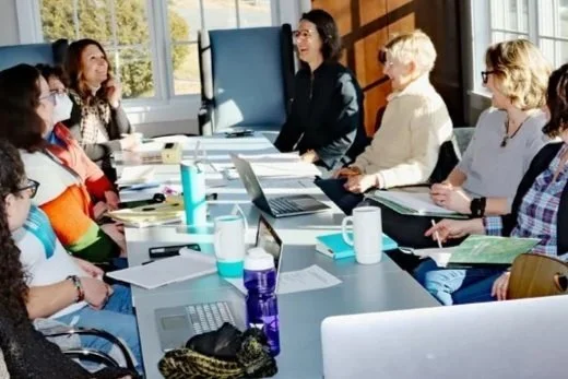 A group of women sit around a table with papers, computers, and travel mugs in front of them. They are smiling and laughing with light coming in through the windows behind them.
