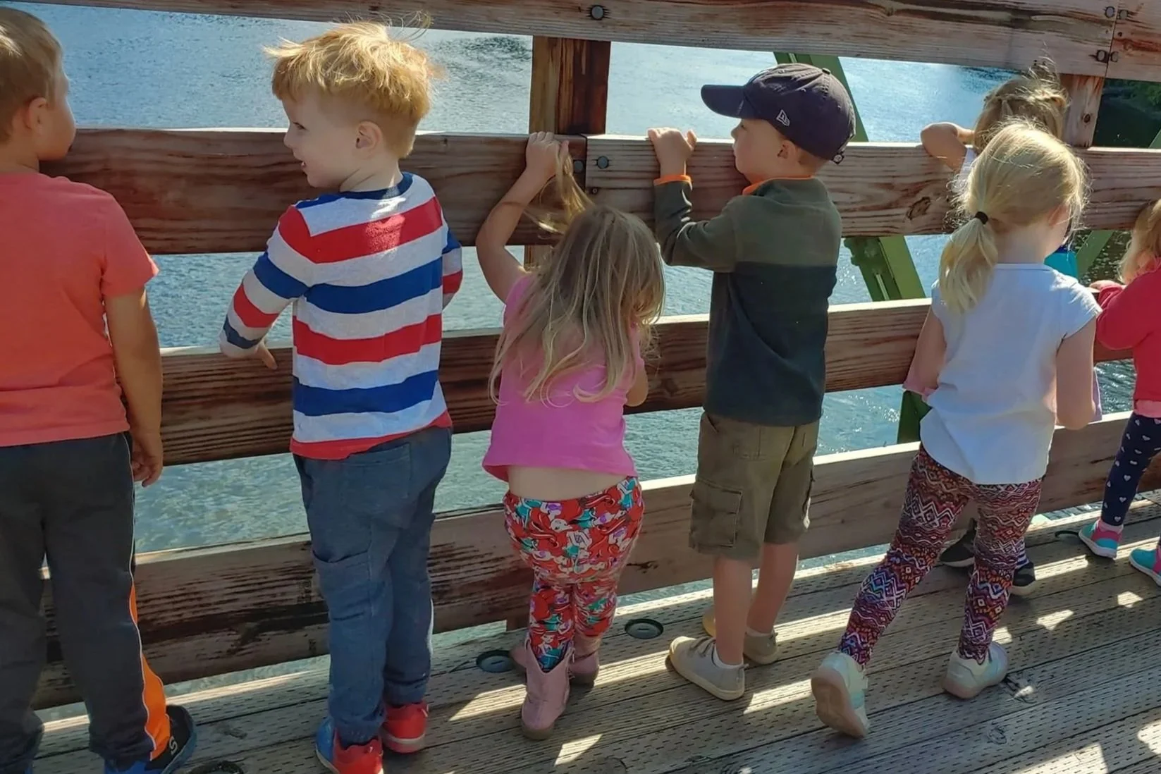 A group of children stand looking through the slats of a bridge railing at the water below on a sunny day. Two of the children are looking at each other and smiling.