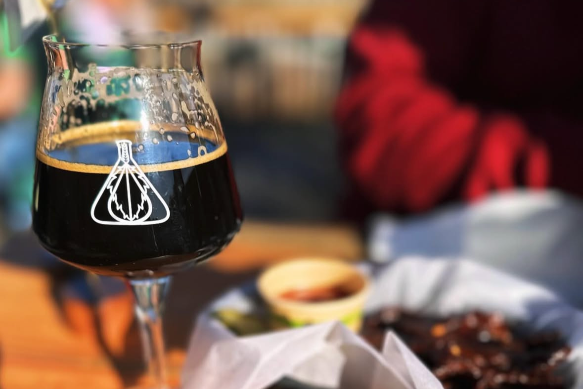 A stem glass with a dark beer and logo from Bound By Fate is in the foreground.  In the background out of focus there is a plate of food and dipping sauce on a wooden table.