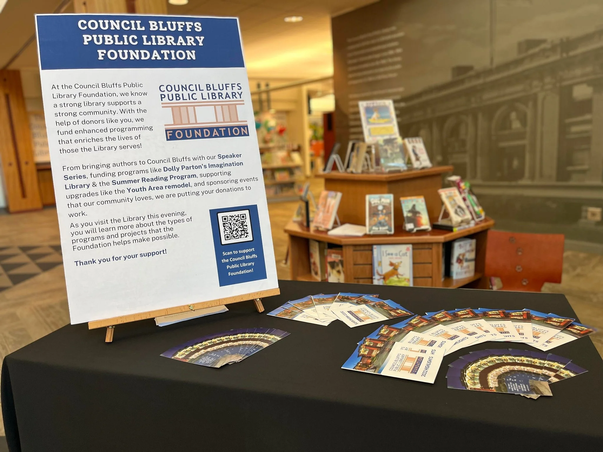 Display for the Library Foundation. There is a poster that says "Council Bluffs Public Library Foundation" and an array of materials on a black tablecloth. Behind it is a stand of books.