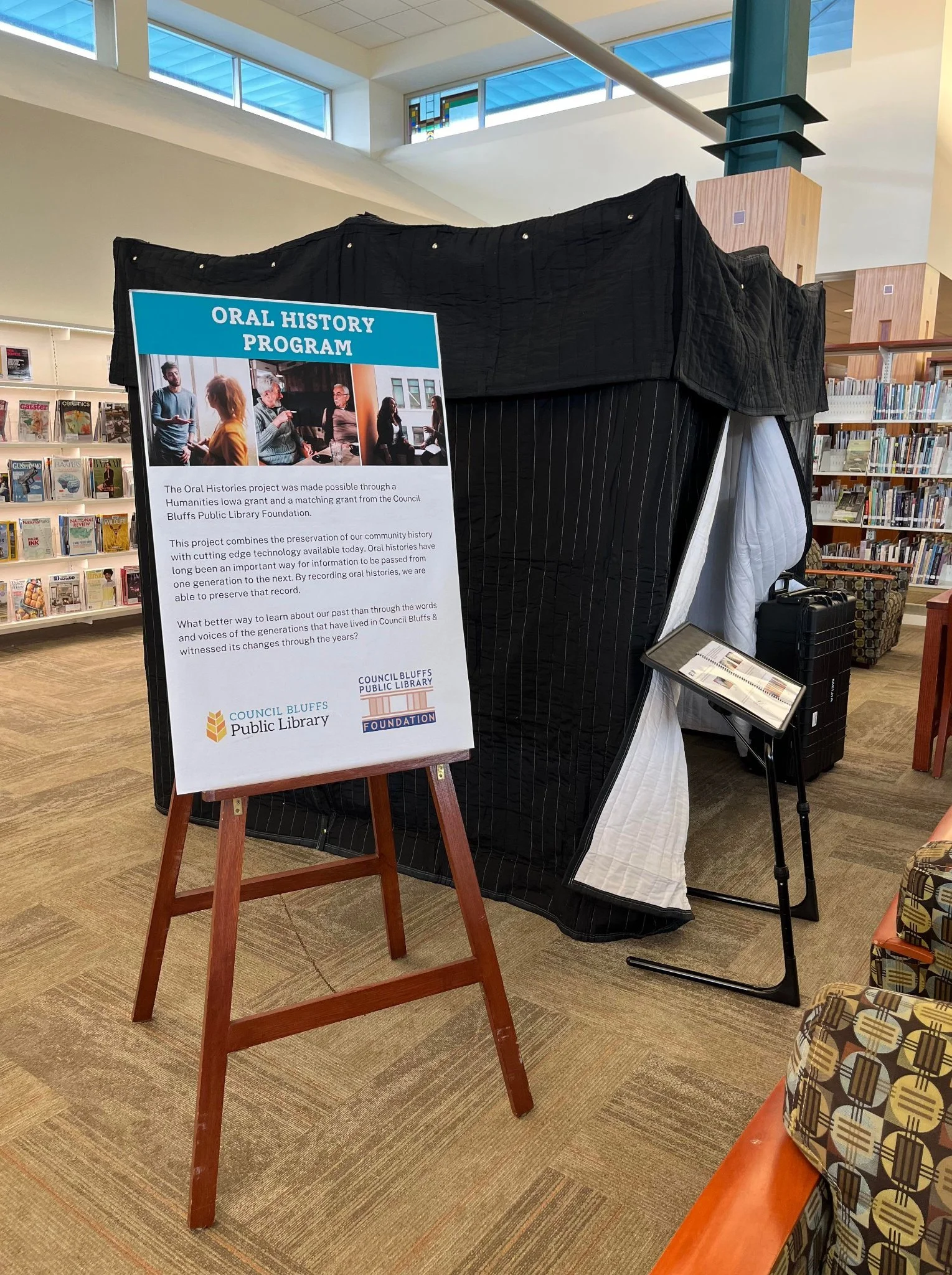 Image of a poster, with the words "oral History Program" in front of a tent (for recording. The tent is set up inside of a large library building.