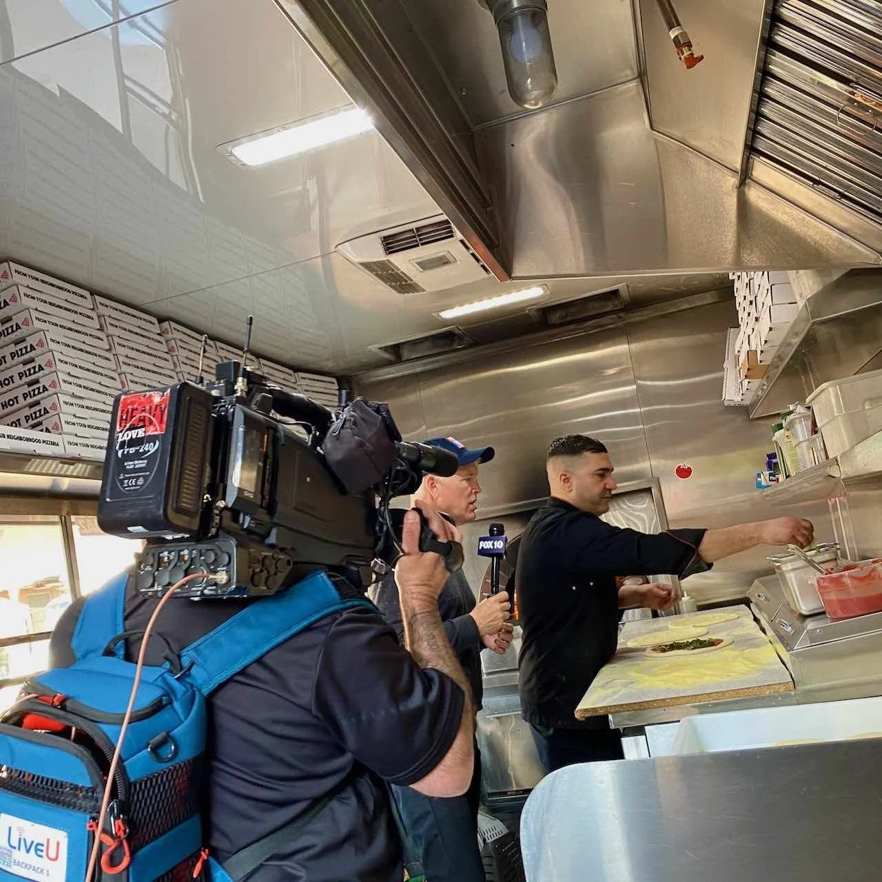 FOX 10’s Cory McCloskey and cameraman film Oualid Ferhati making pizza inside the Re Di Roma food truck during live broadcast.