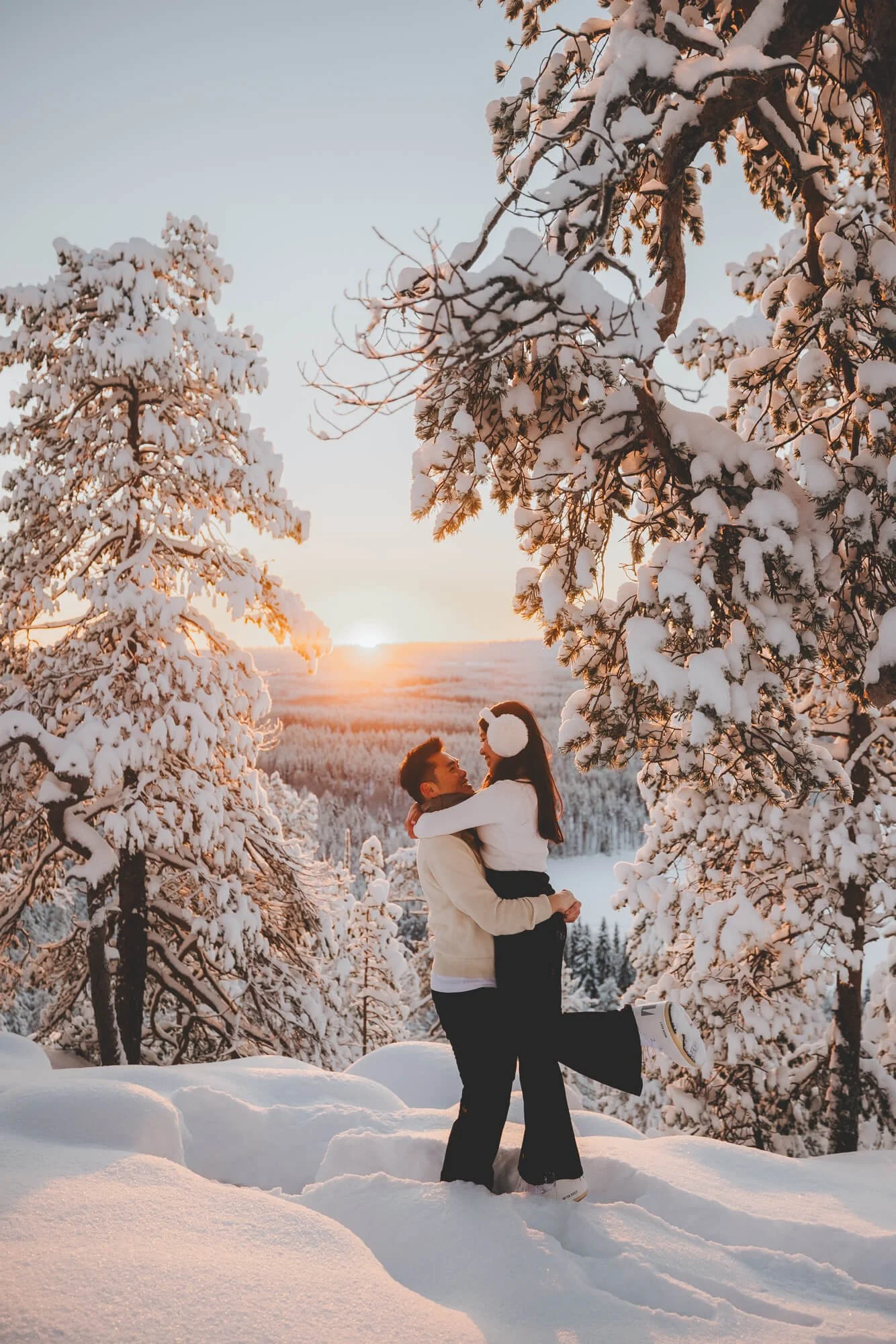 A couple is standing in a snowy landscape, with the man lifting the woman off the ground as they face each other and smile. They are surrounded by snow-covered trees, and the sun is setting in the background, casting a warm glow over the scene.