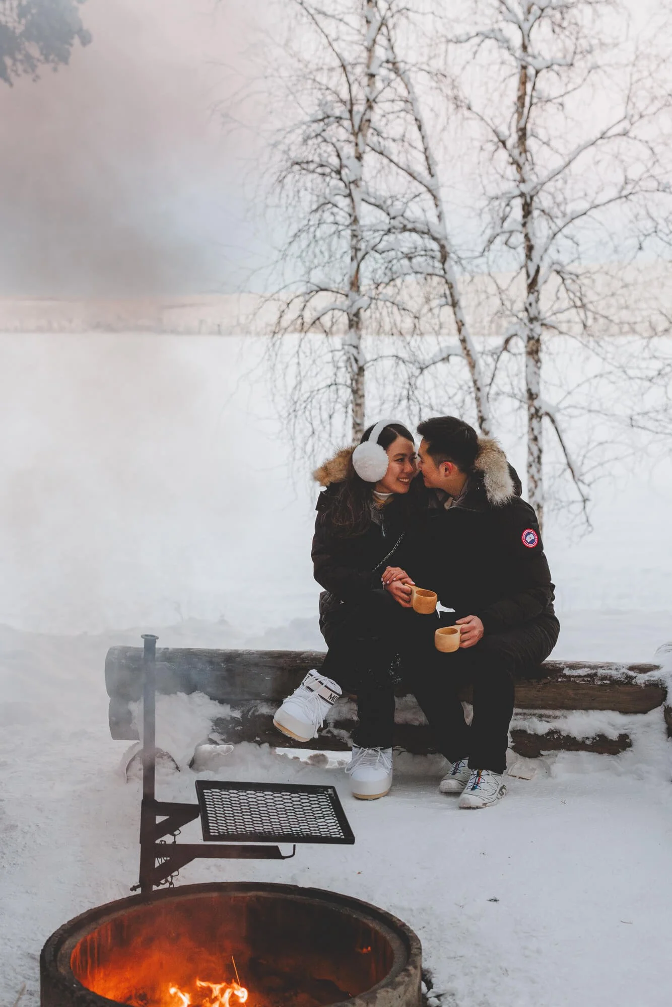 Couple photos in Rovaniemi and Lapland. A couple sitting outdoors on a snow-covered bench, holding mugs, with a fire pit in front of them. They are smiling and leaning close to each other, dressed warmly, with snow-covered trees in the background.