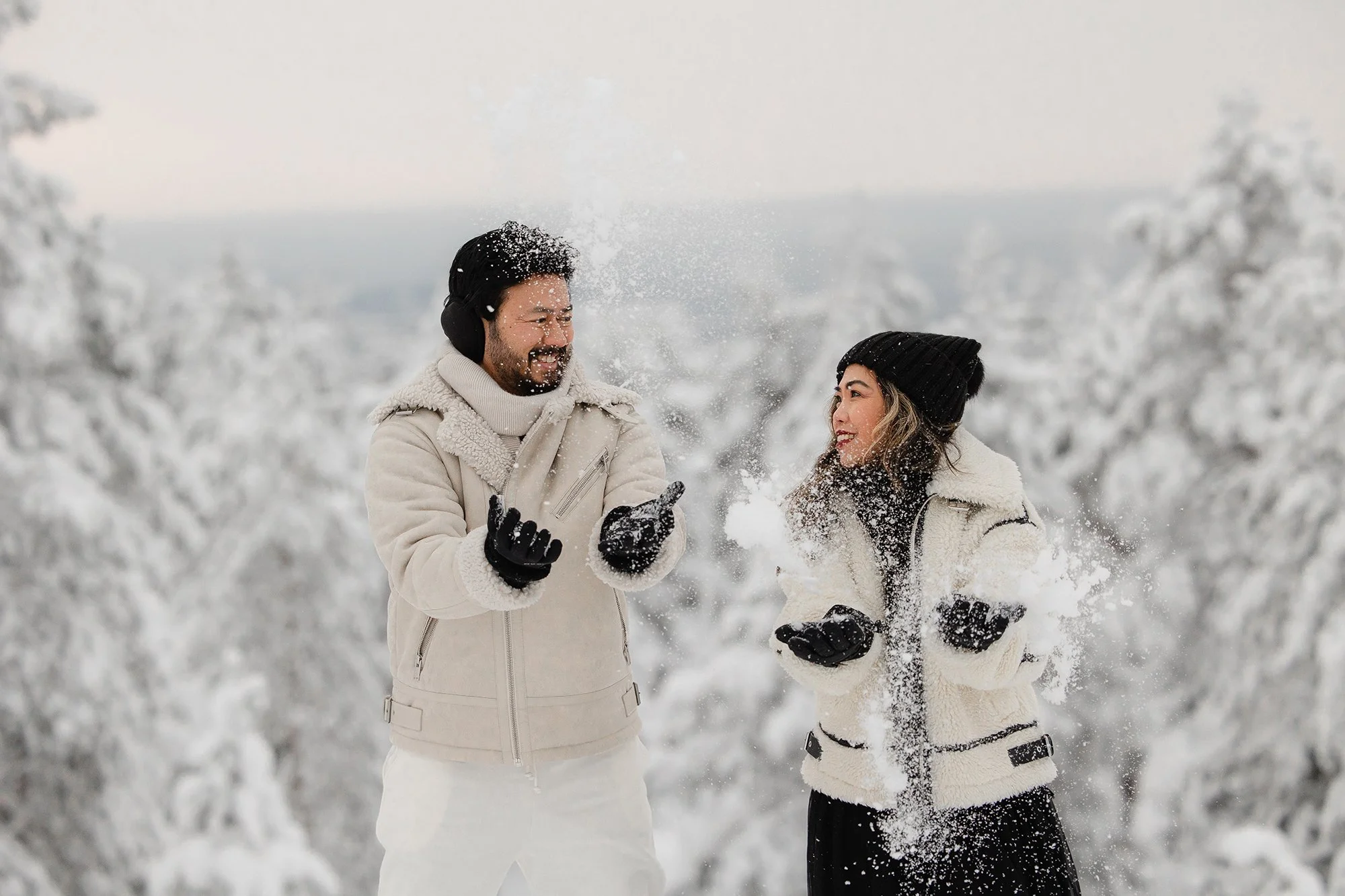 Photographer in Lapland. A man and woman smiling and playing in the snow in a winter landscape with snow-covered trees, wearing winter jackets, gloves, and hats.