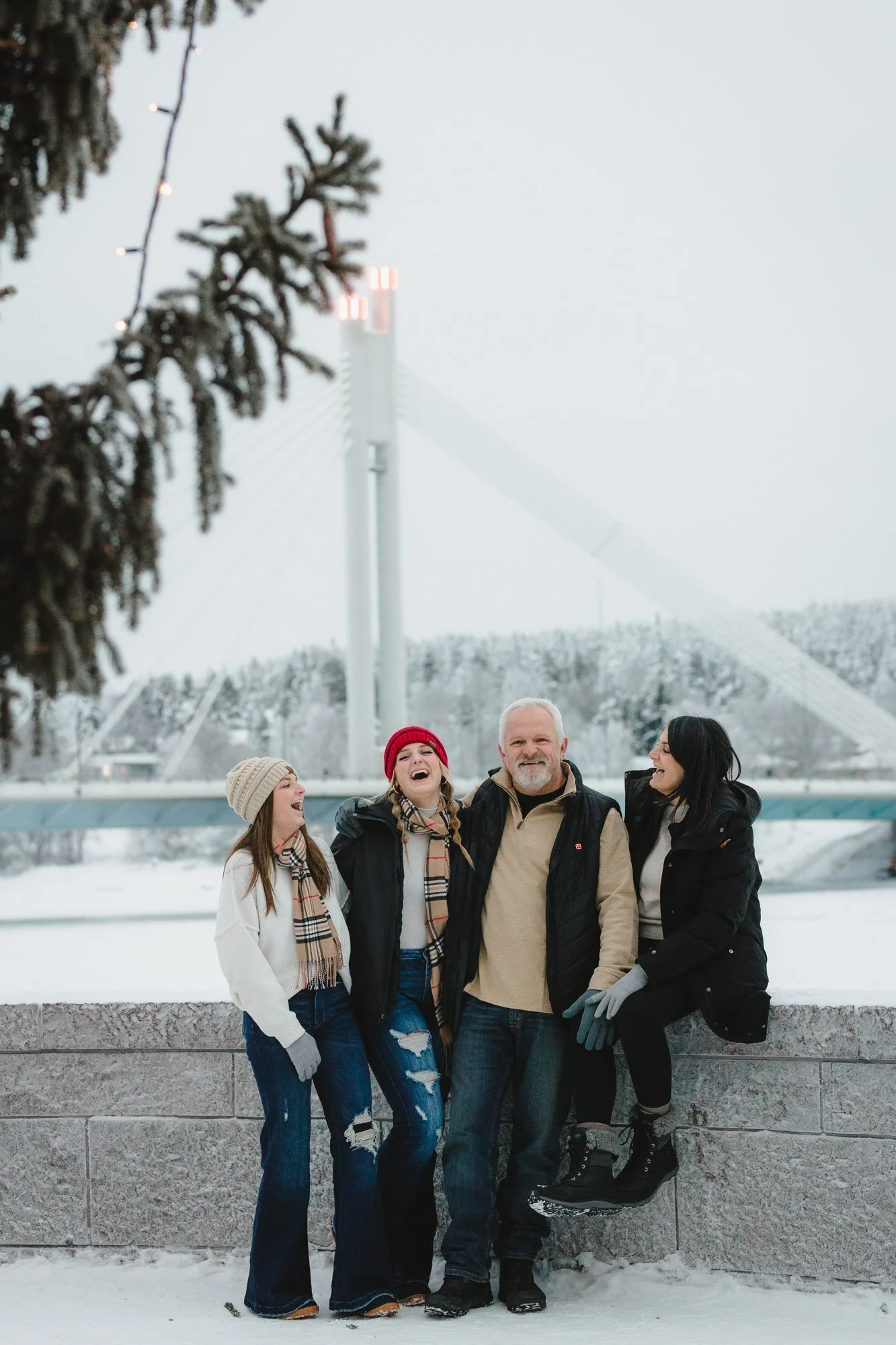 A group of five people posing outdoors in winter snow, standing in front of a stone barrier, with a snow-covered landscape, trees, and a bridge in the background. One person is lifted off the ground, supported by others, and they are all smiling and 