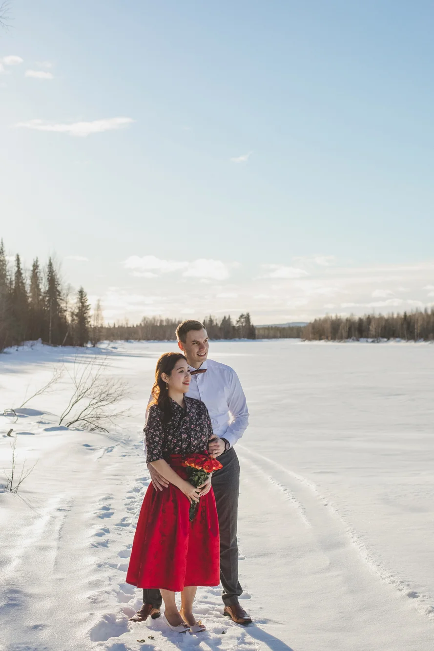 A couple stands on a snow-covered path near a frozen lake, holding a bouquet of red flowers, with a forest in the background and a clear blue sky.