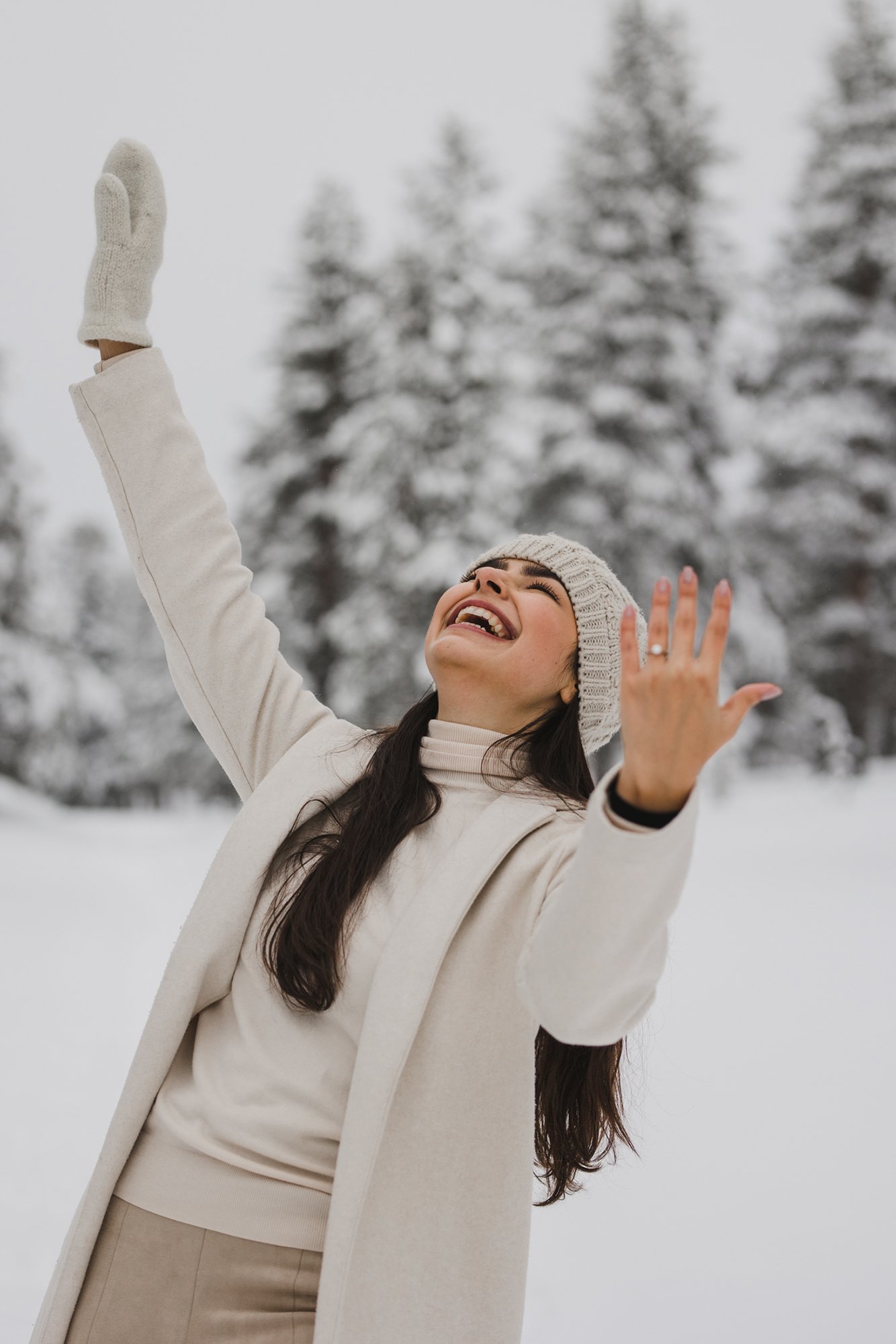 A woman smiling and raising her arms in a snowy outdoor setting with snow-covered trees in the background.