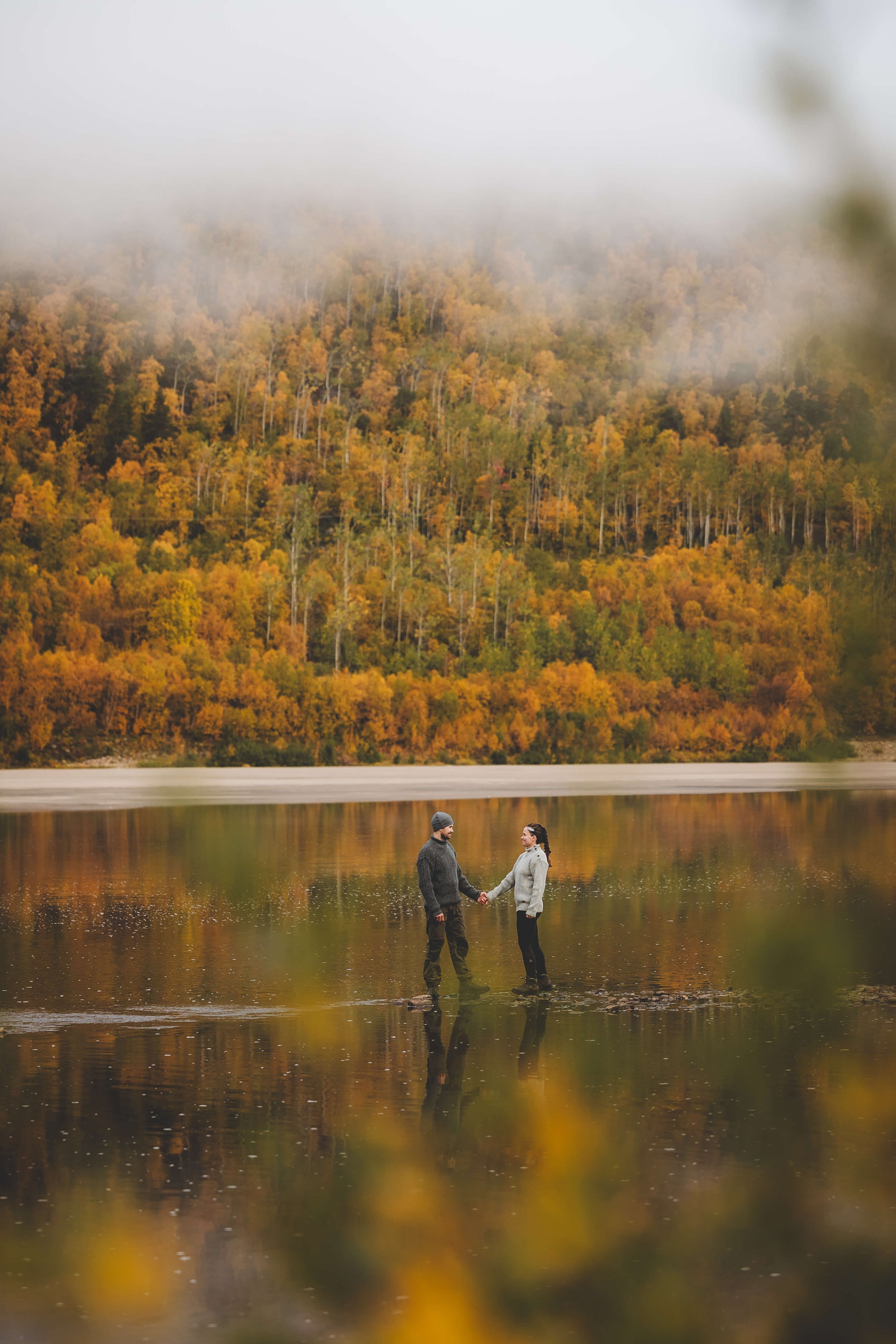 A couple holding hands and standing in a calm lake with a backdrop of colorful autumn trees and fog on a mountain.