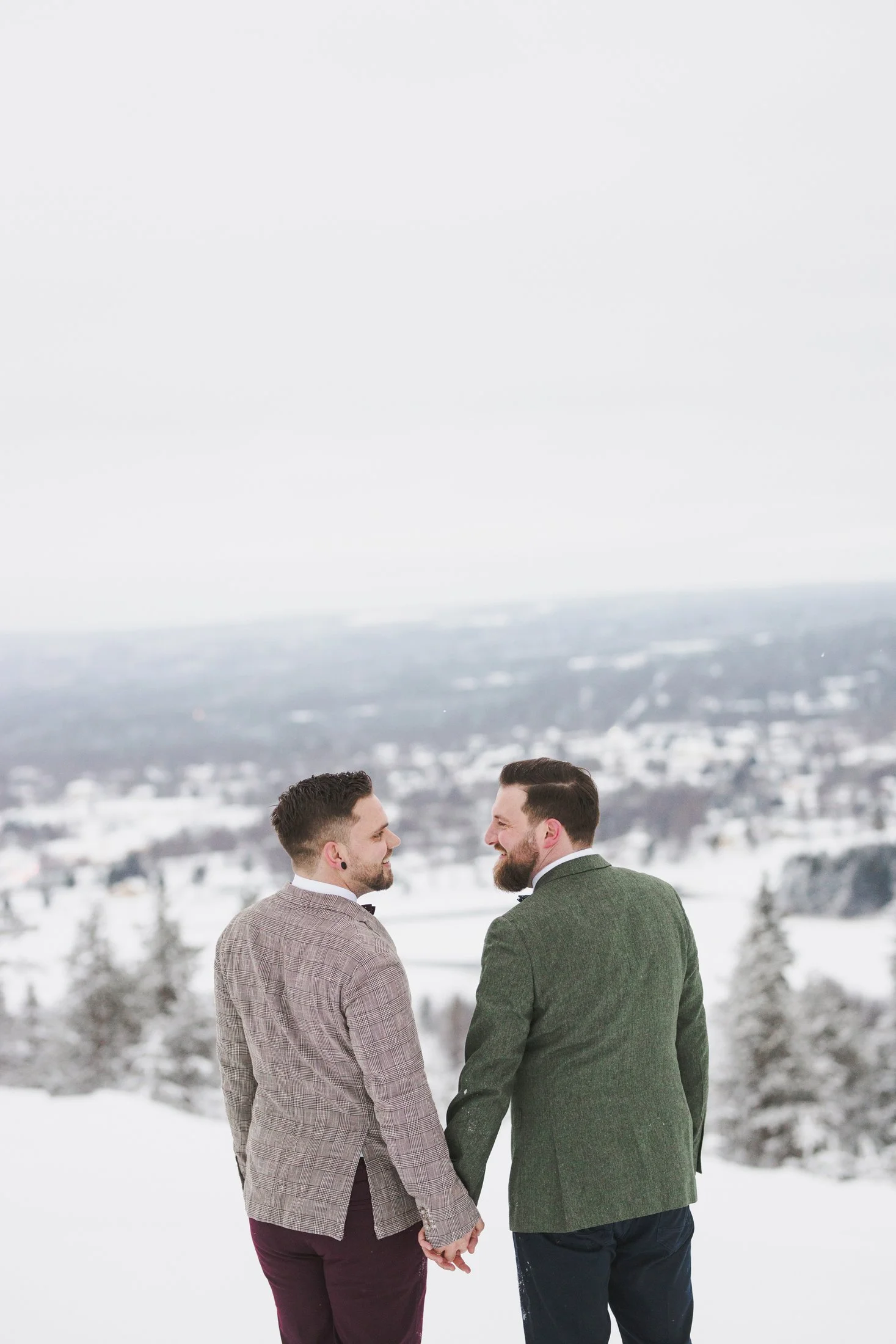 Two men with beards in suits holding hands and smiling at each other outdoors in a snowy landscape with snow-covered trees and hills in the background. Lapland wedding photos. Gay wedding.