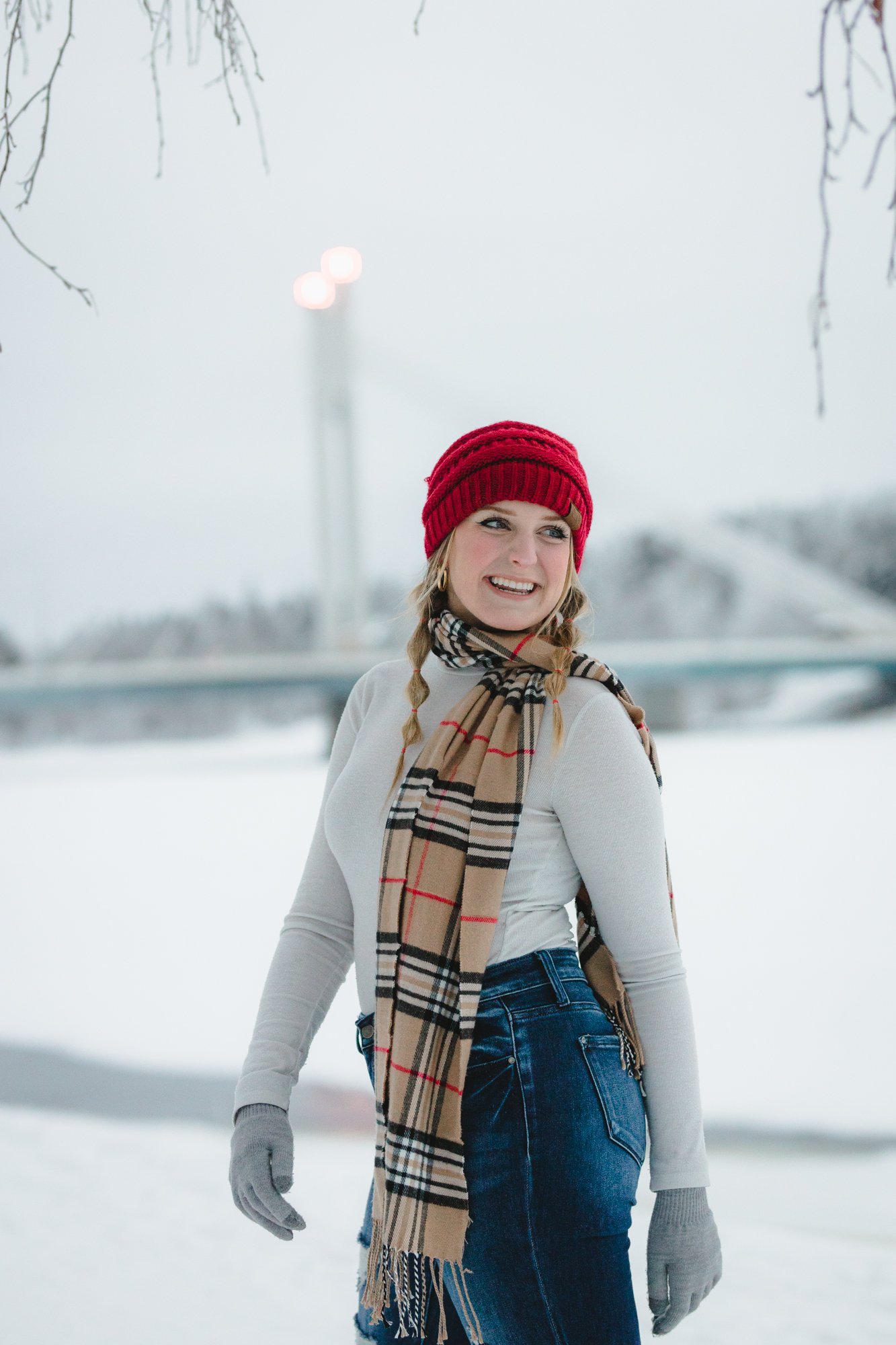 A young woman in a red knit hat, plaid scarf, white long sleeve shirt, and blue jeans standing outdoors in a snowy landscape with trees and a bridge, smiling and looking to the side.