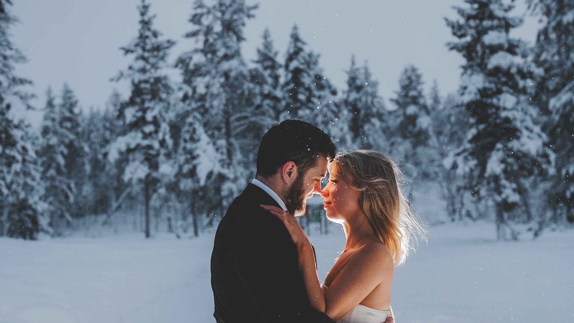 Photographer in Rovaniemi. A couple embracing each other closely in a snowy outdoor setting with snow-covered trees in the background. The man is dressed in a dark suit, and the woman appears to be wearing a light-colored dress with her hair down.  