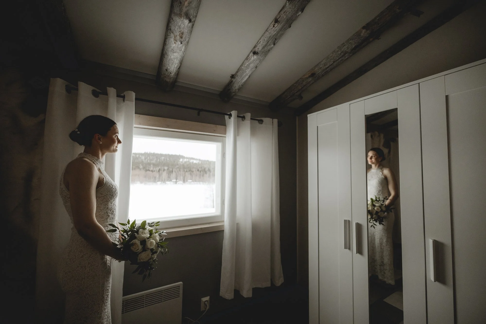 A woman in a lace dress holding a bouquet of white roses standing near a window with white curtains, looking at her reflection in a mirror on a white wardrobe in a rustic room with exposed wooden beams on the ceiling. Wedding photography Lapland.