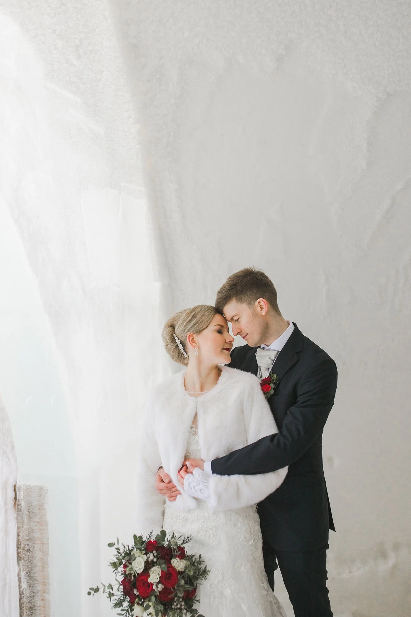 A bride and groom share an intimate moment during their wedding. The bride wears a white lace wedding dress and a fur stole, holding a bouquet of white and red roses. Wedding photoshoot in Rovaniemi and Lapland.