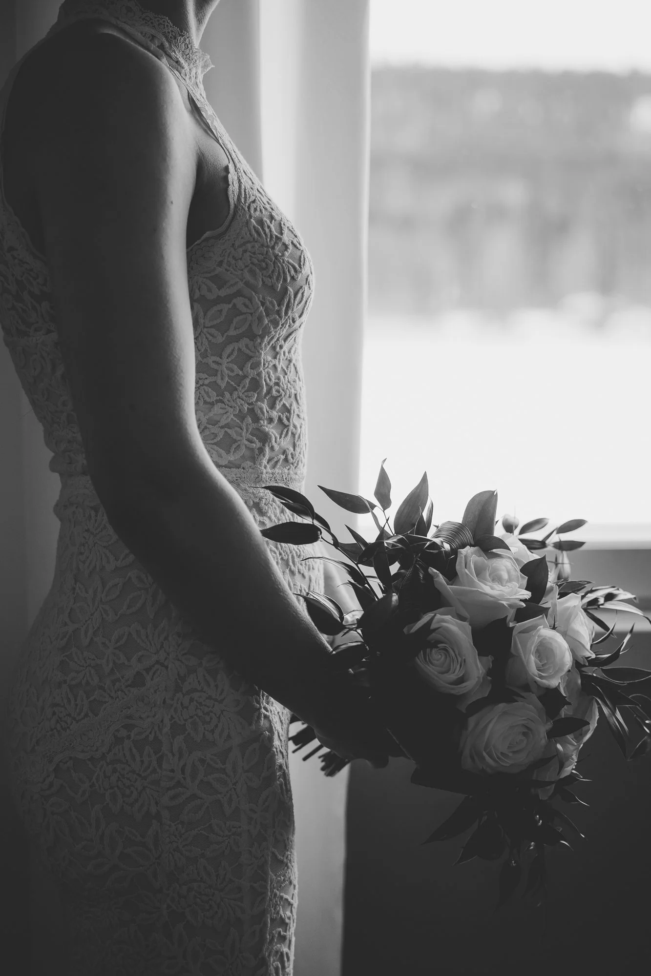 A black and white photograph of a woman in a lace dress holding a bouquet of roses, standing by a window with natural light.