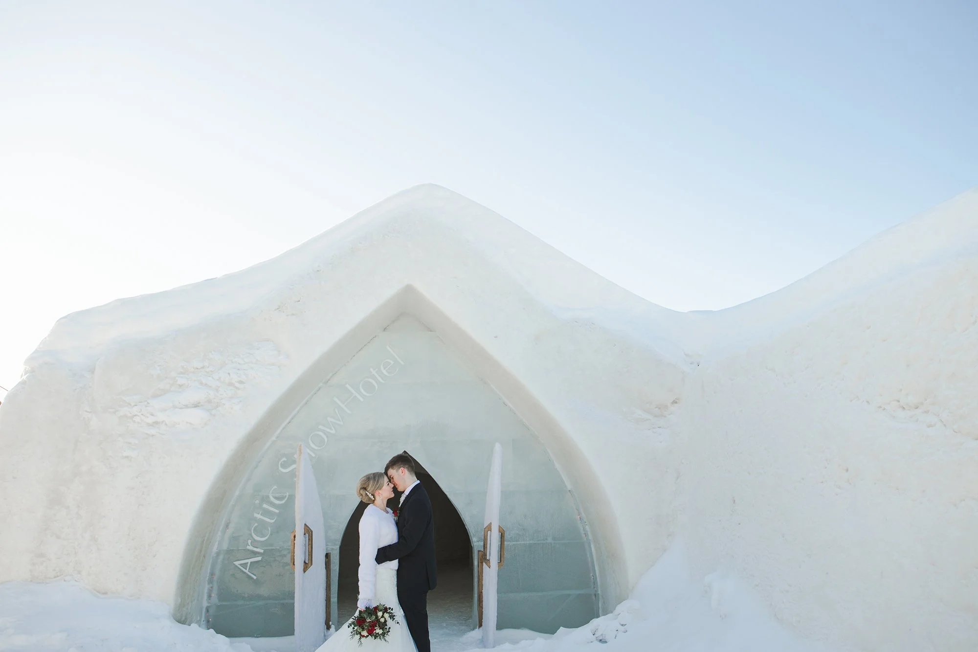 Wedding photoshoot in Lapland. A bride and groom standing close together inside an ice hotel, holding hands and looking at each other, with snowy mountain peaks visible in the background.