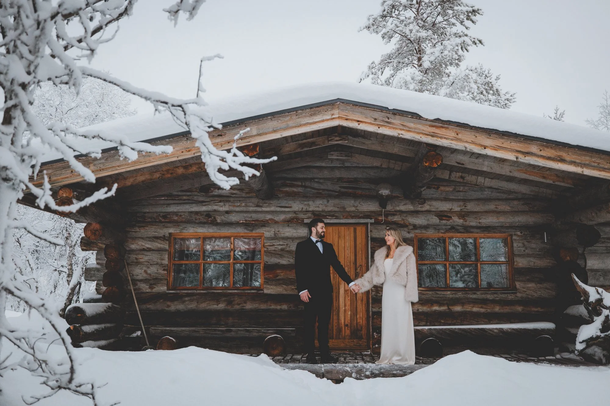 A couple dressed in wedding attire standing outside a rustic log cabin in a snowy landscape, holding hands and looking at each other.