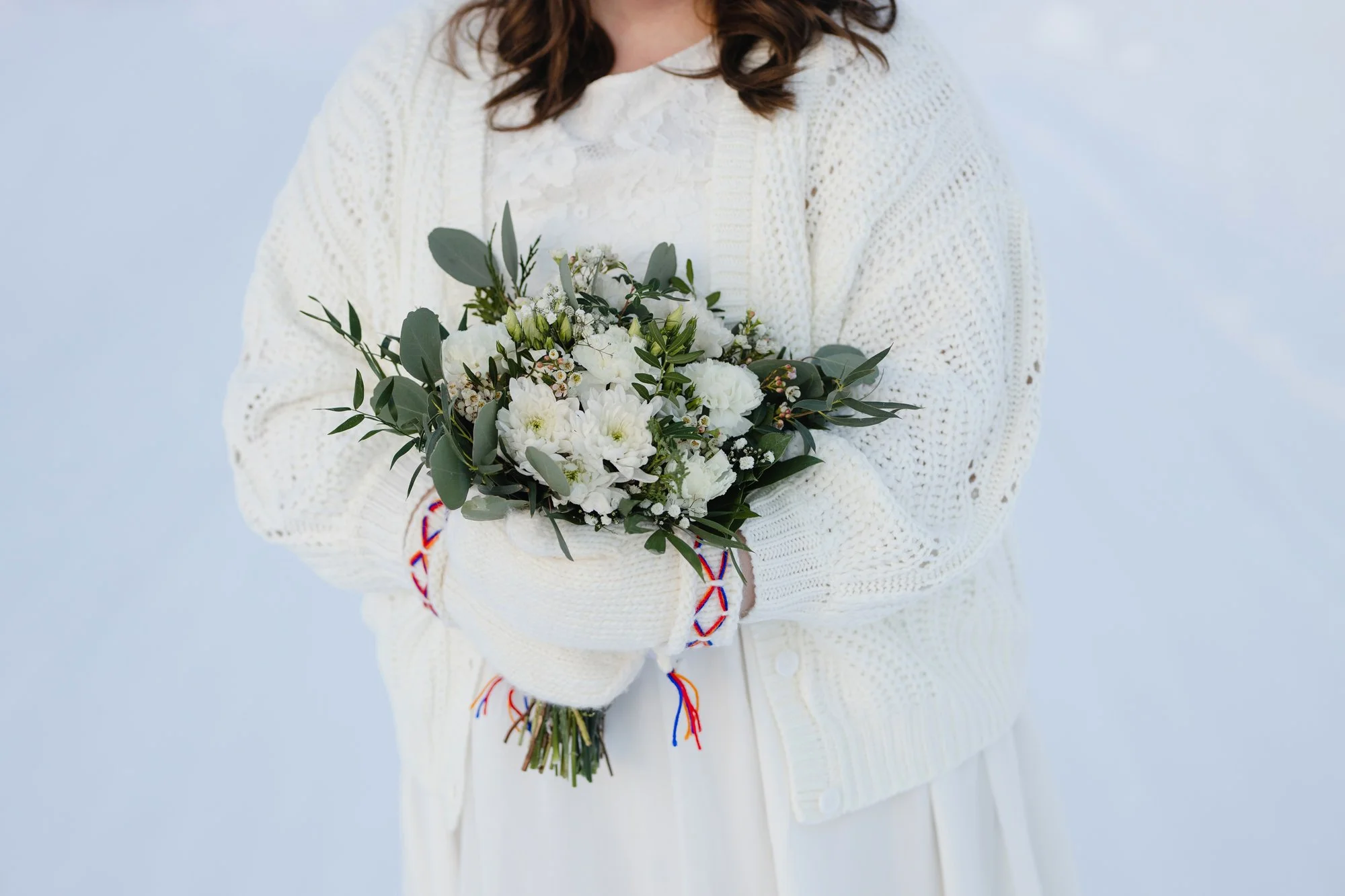 A bride in Lapland wearing a white knit sweater and white lace dress holding a bouquet of white flowers and greenery.