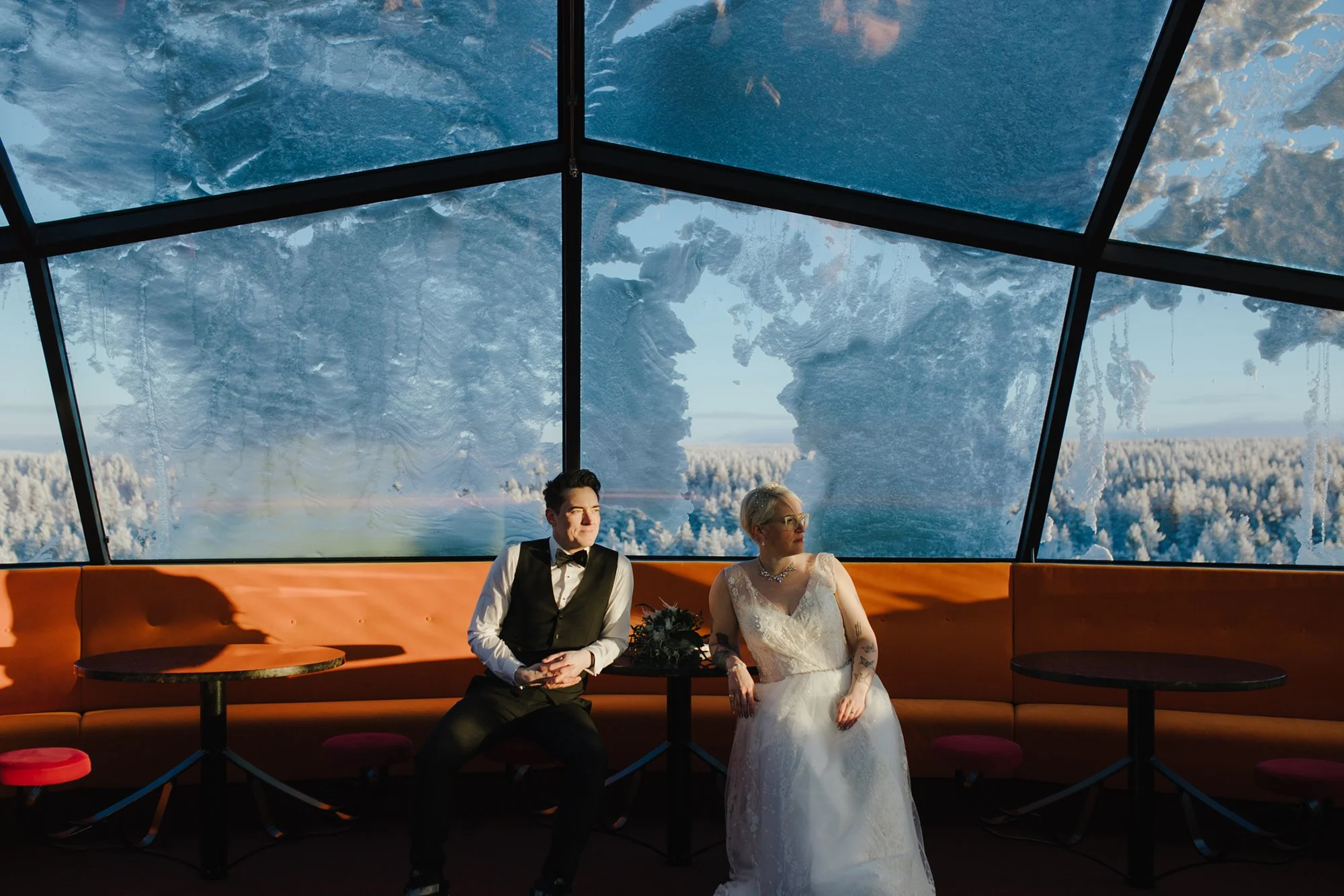A couple dressed in wedding attire sitting on an orange bench inside a building with large glass windows showing snow-covered trees and mountains outside. Tina the Lapland Photographer shoots amazing wedding photos in Rovaniemi and whole Lapland.