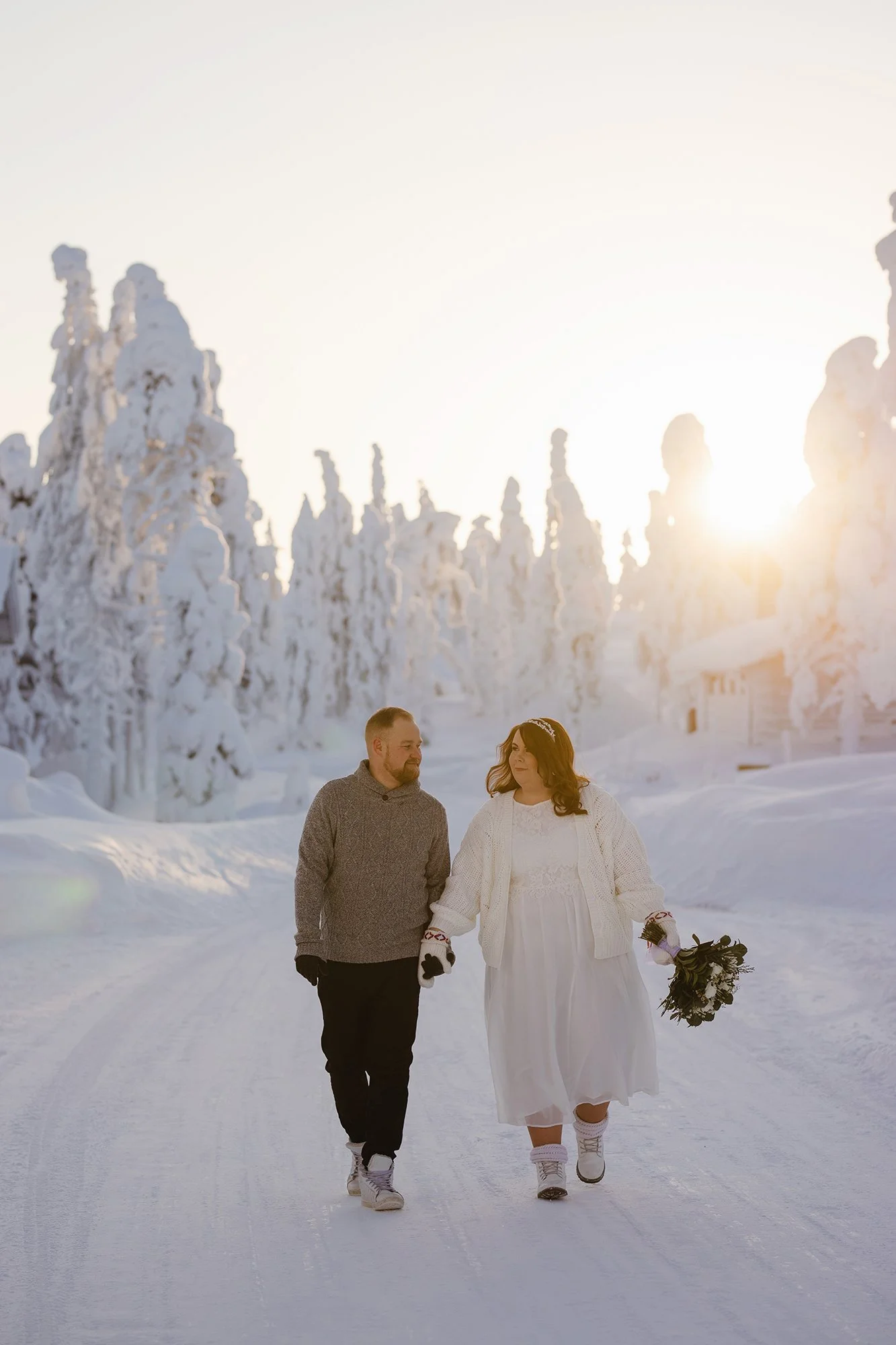 A couple walking in a snowy landscape with snow-covered trees and the sun setting behind them, the woman wearing a white dress and holding a bouquet. Tina the Lapland Photographer. Wedding photos.