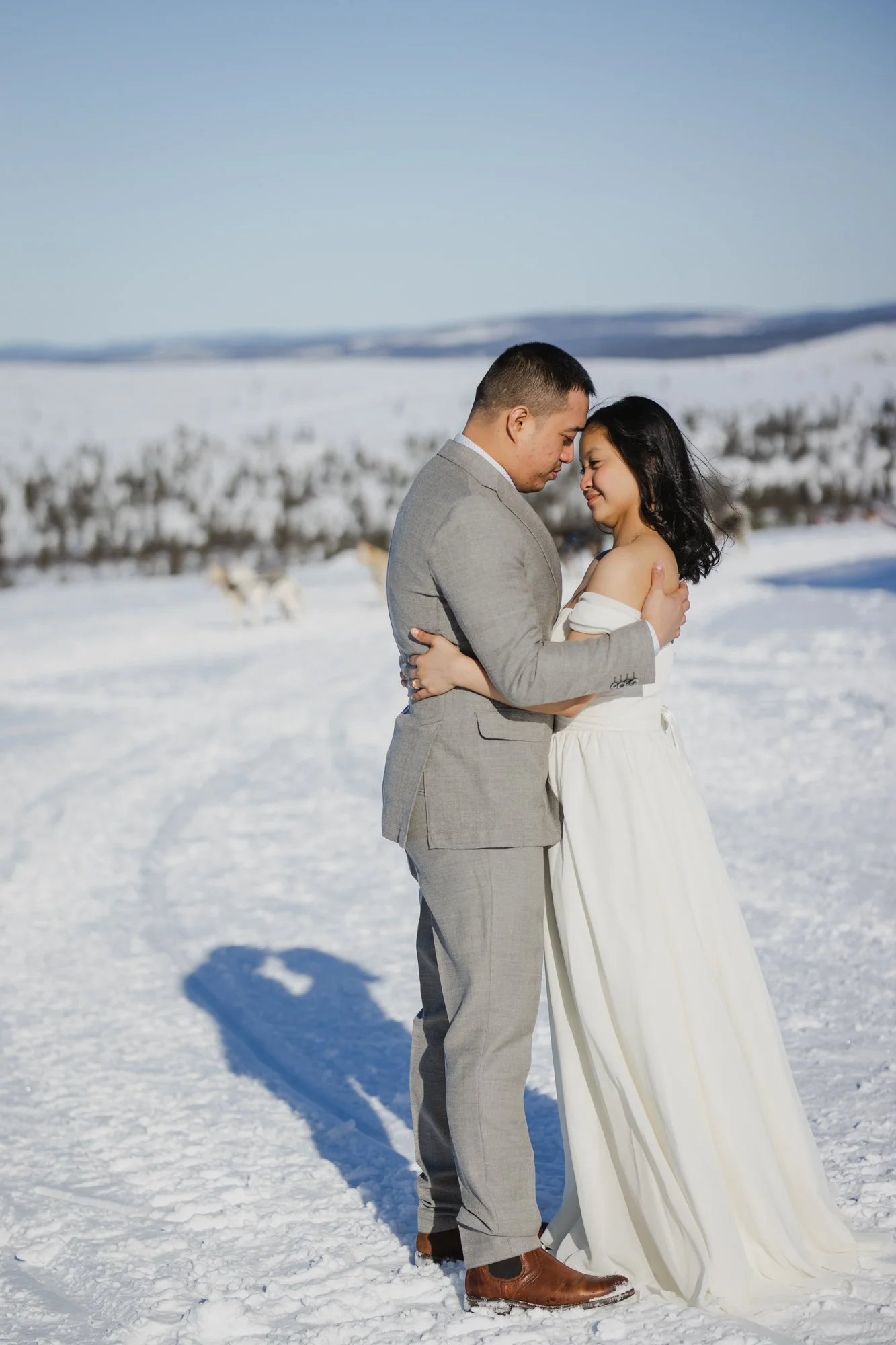 Wedding photoshoot in Lapland. A couple dressed in wedding attire standing close together in a snowy landscape, holding each other with their foreheads touching and eyes closed, with mountains and trees in the background.