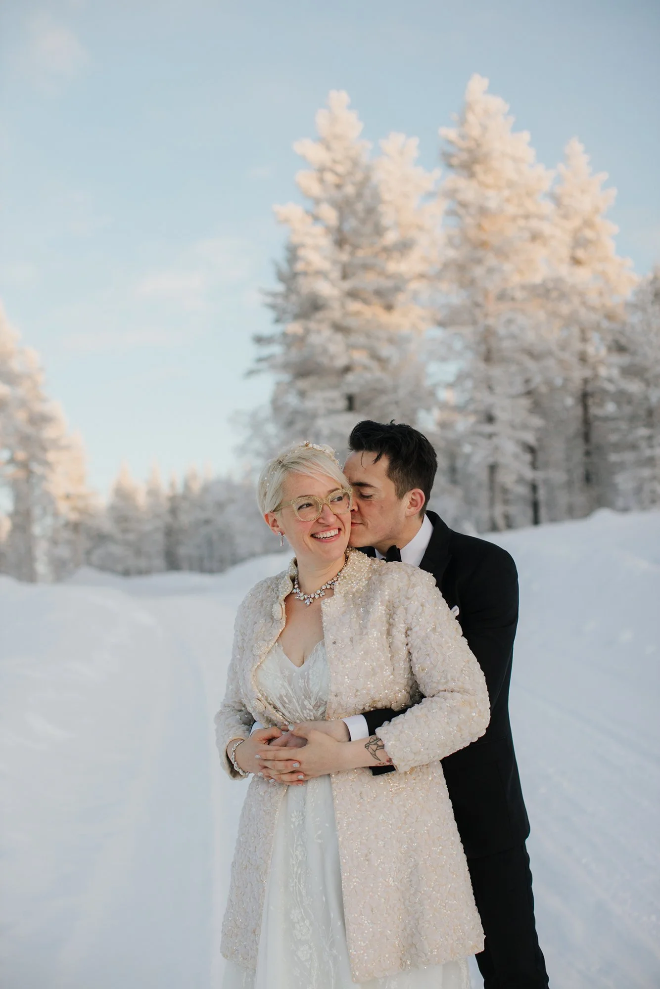 A smiling woman in a wedding dress and pearls, with glasses, is being kissed on the cheek by a man in a tuxedo, in a snowy forest with snow-covered trees in the background. Wedding photoshoot in Lapland.