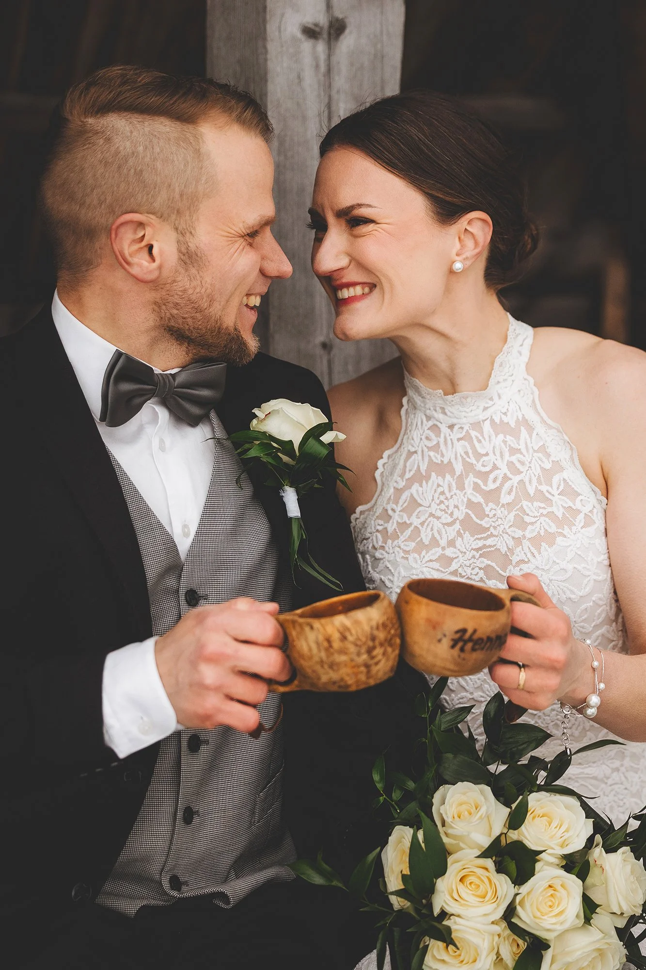 A smiling bride and groom holding wooden cups during their wedding celebration, with the bride in a lace dress and the groom in a tuxedo, surrounded by white roses.