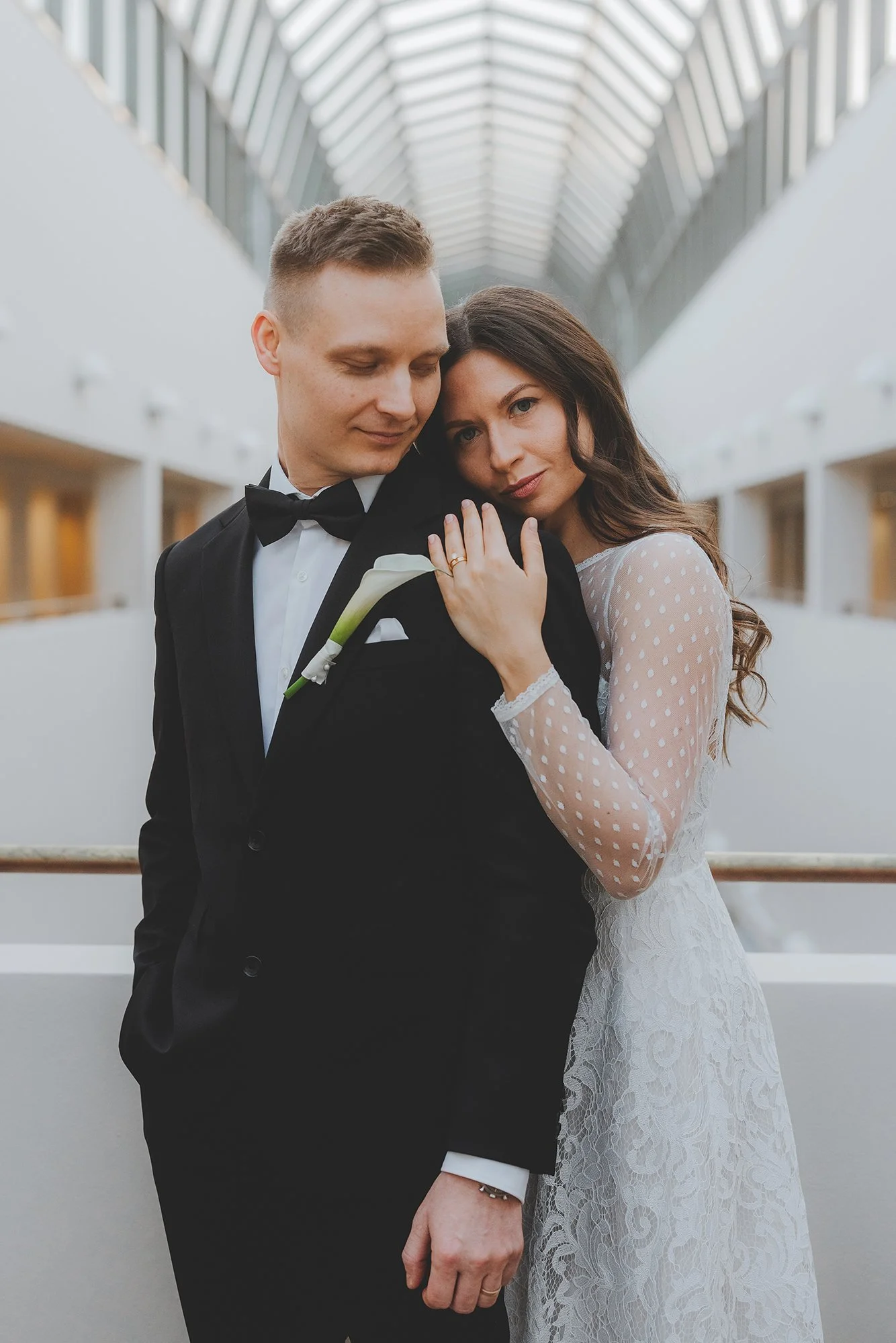 A bride and groom in formal wedding attire embrace on a modern indoor walkway with a glass ceiling.