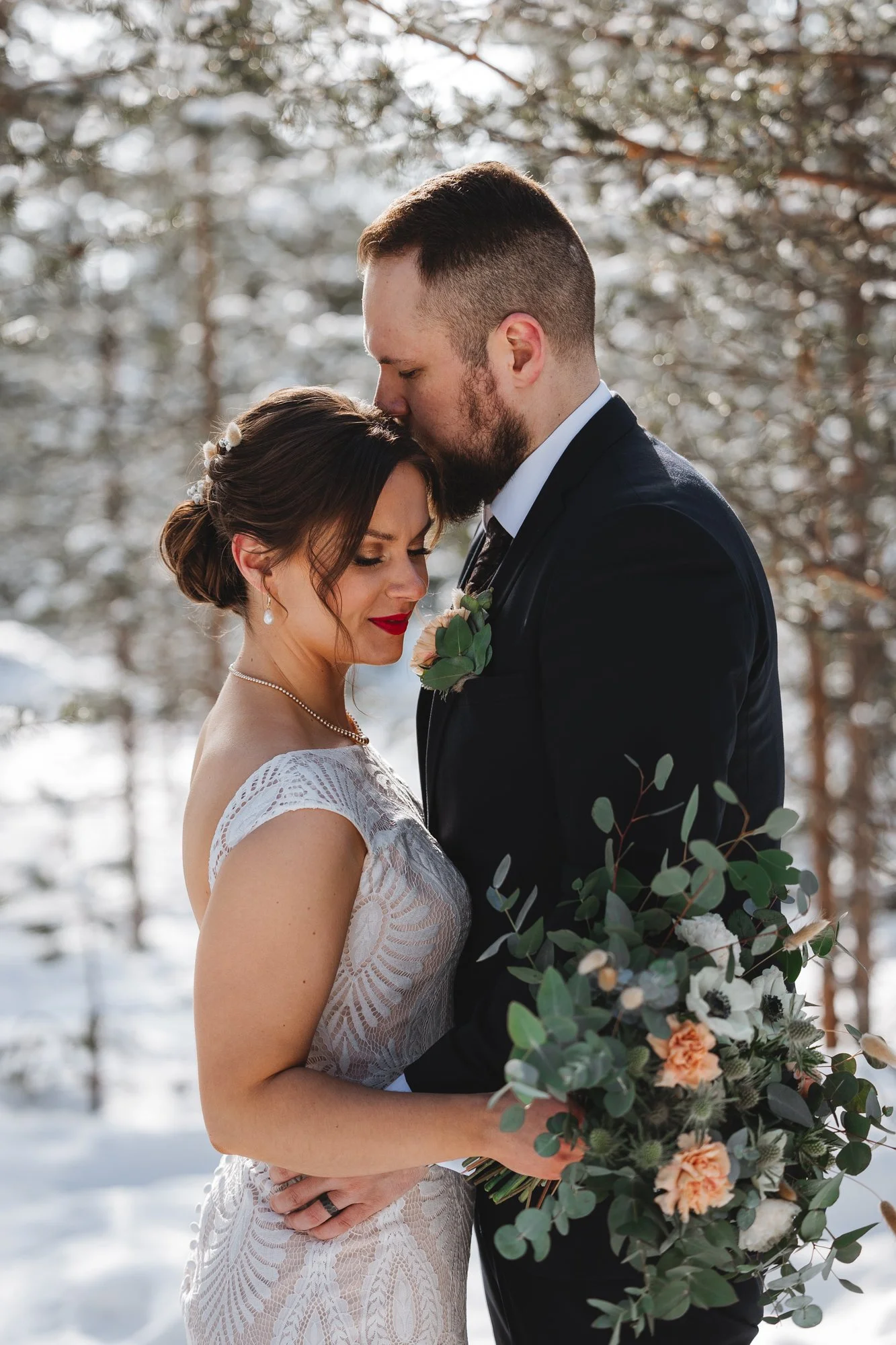 A bride and groom embracing outdoors during winter. The bride wears a lace wedding dress and has dark hair styled in an updo with pearl accessories. The groom wears a black suit and has a beard. They are holding a bouquet of flowers and greenery, sta