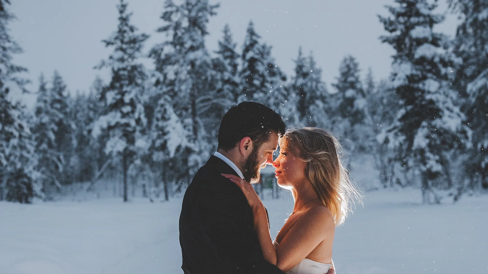 A couple dressed formally, embracing tenderly in a snowy forest during winter, with snow-covered trees in the background. Tina the Lapland Photographer.