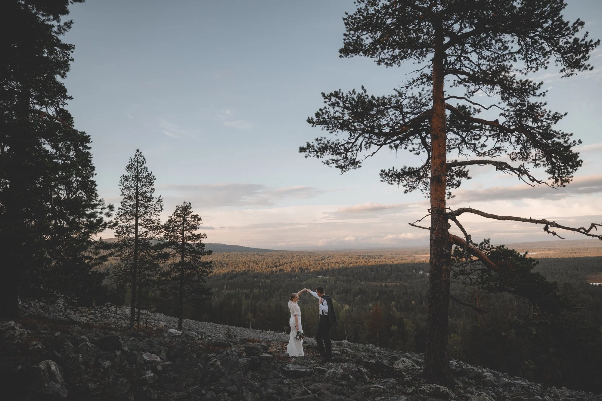 A couple in wedding attire standing on rocky terrain in a scenic outdoor location. They are holding hands and the groom is holding a bouquet of flowers. Tall trees and a vast landscape with forest and sky are in the background.