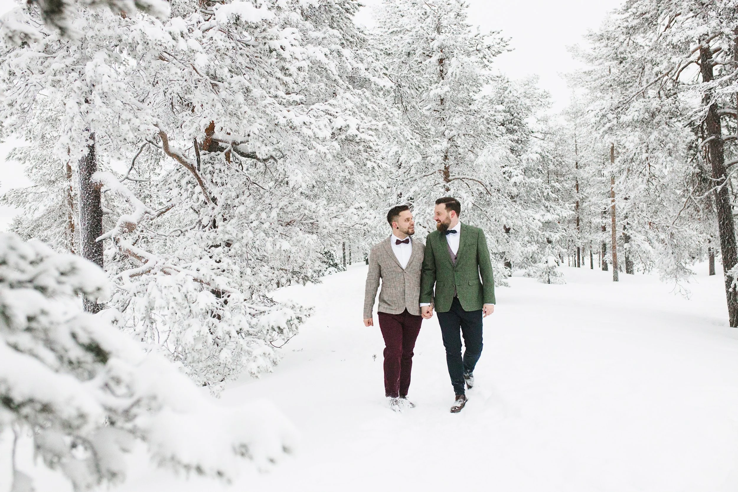 Two men walking hand-in-hand through a snowy forest, dressed in formal suits with bow ties, amidst snow-covered trees on a winter day. Wedding photos for men in Lapland.