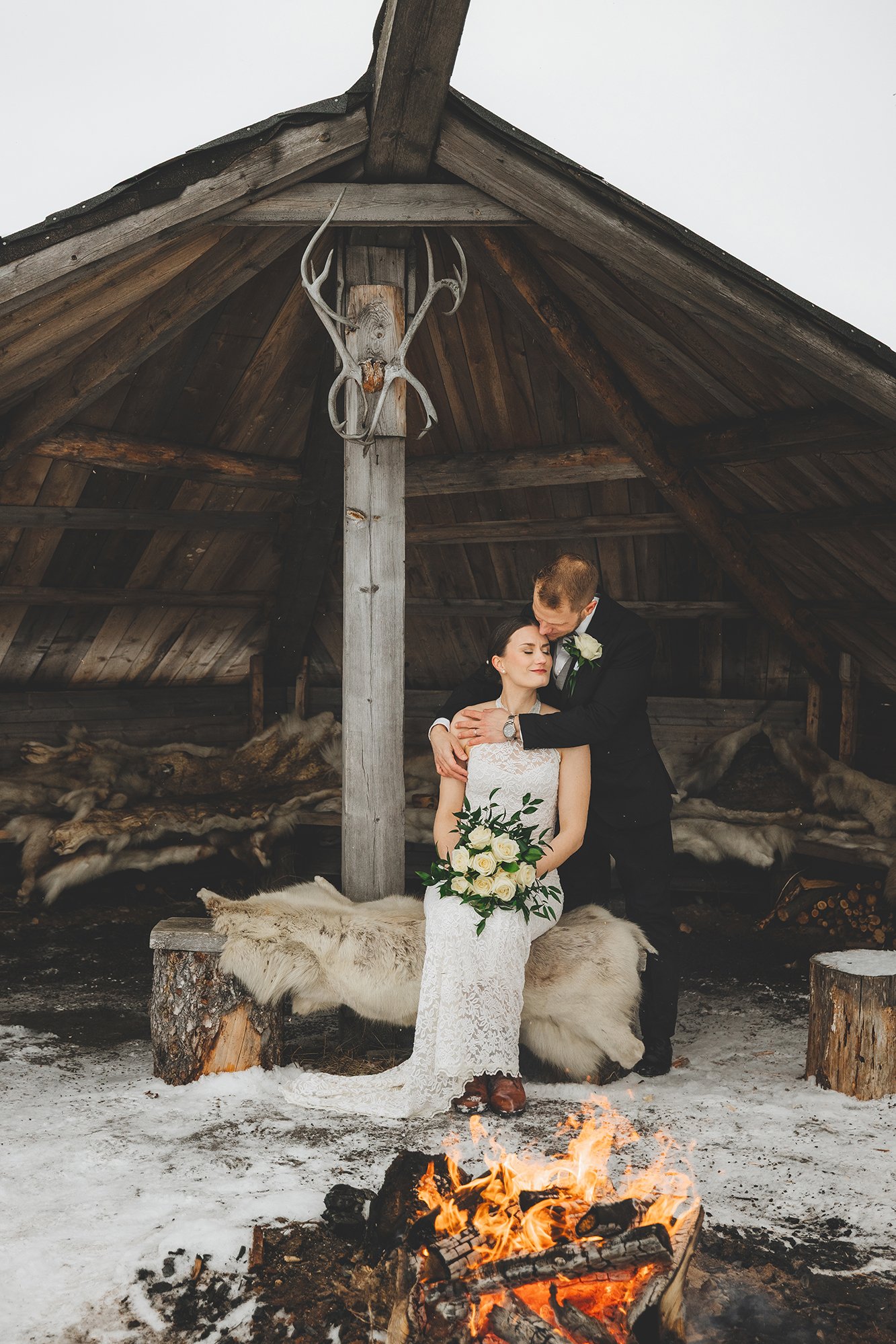 A newly married couple, a woman in a white lace wedding dress holding a bouquet of white roses, and a man in a black suit, embrace under a rustic wooden shelter with animal pelts and a mounted deer skull. There is a small fire burning in the snow in 