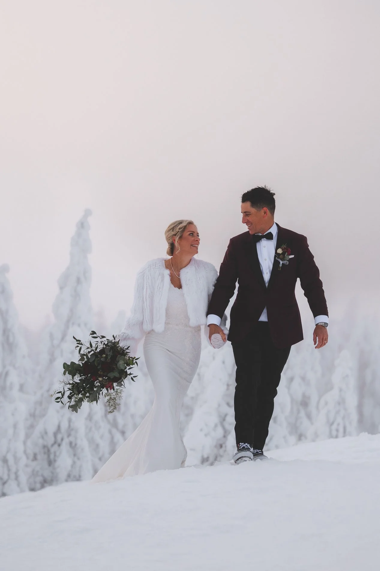 A bride and groom holding hands and smiling in a snowy outdoor setting, with snow-covered trees in the background. Proposal or wedding photos in Finnish Lapland.
