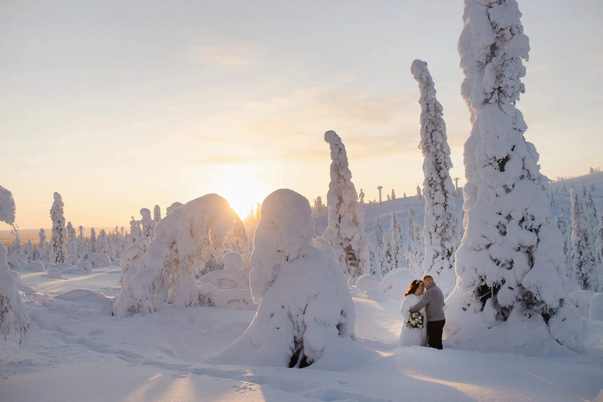Wedding photos in Lapland. A couple stands close together in a snowy landscape, with the woman holding a bouquet of flowers, underneath snow-covered trees at sunset.