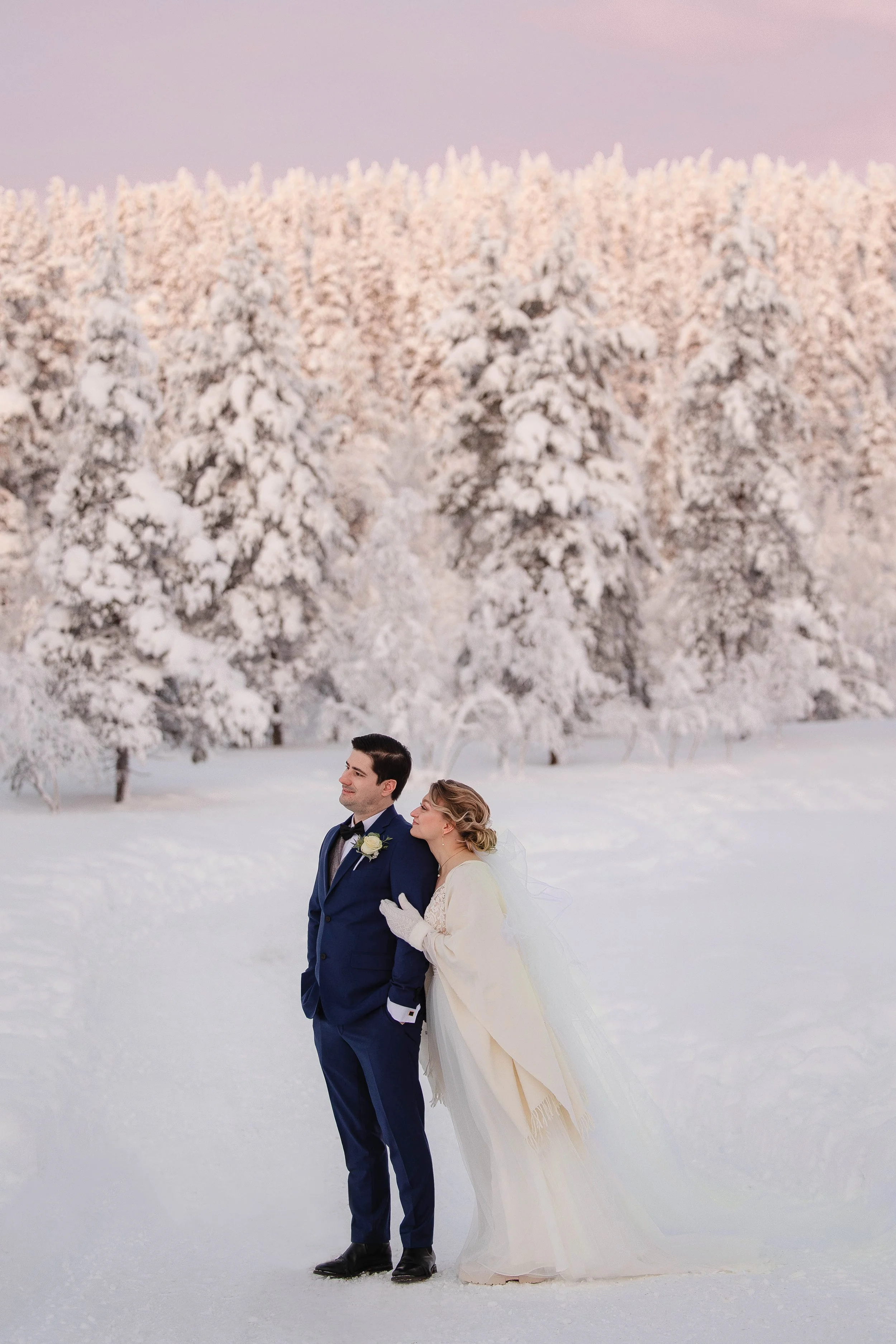 A photo of bride and groom standing together outdoors in a snowy landscape with snow-covered trees in the background. Wedding photos in Rovaniemi, Lapland.