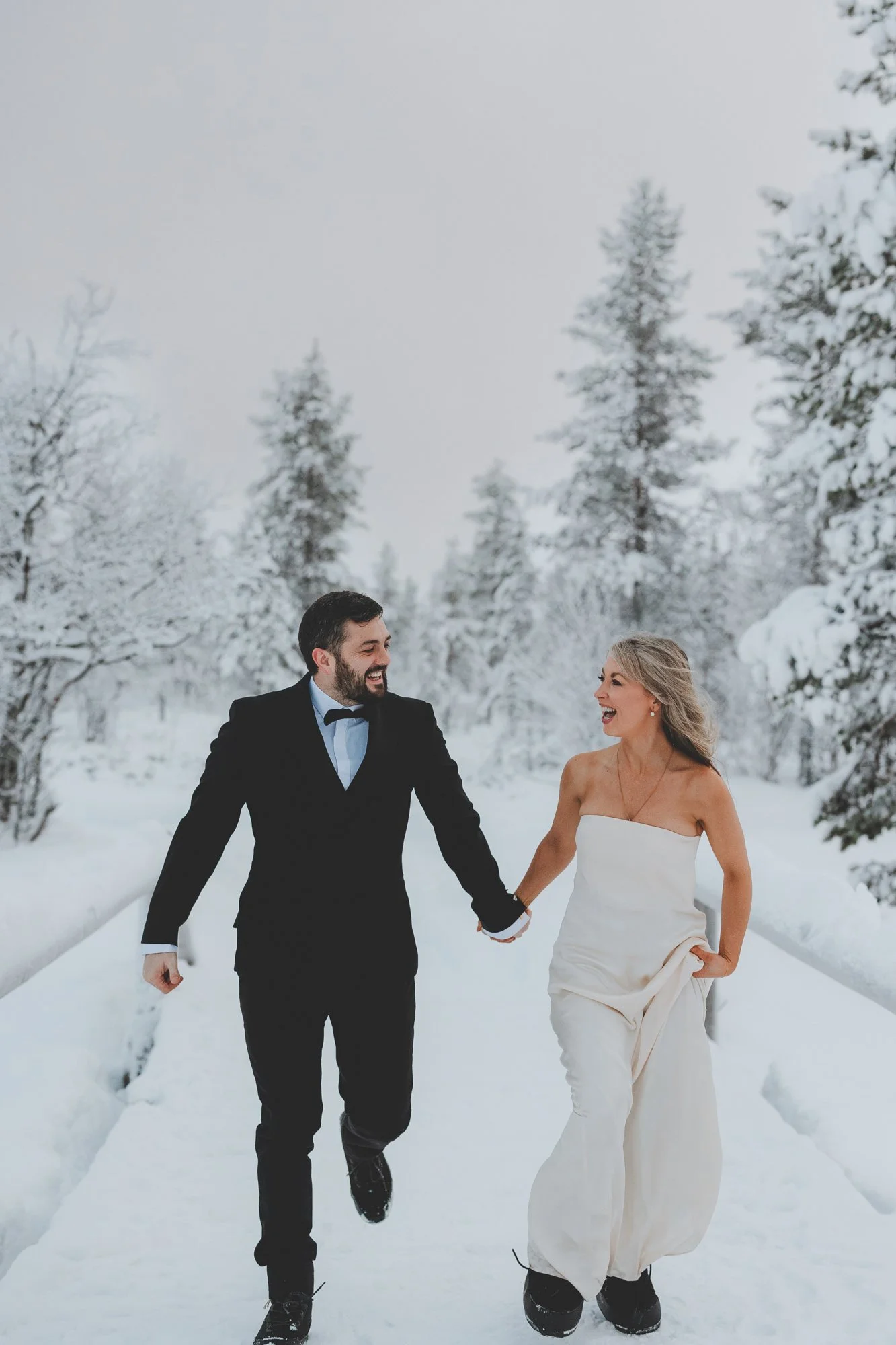 A happy couple holding hands and walking in the snow on a winter day, surrounded by snow-covered trees.