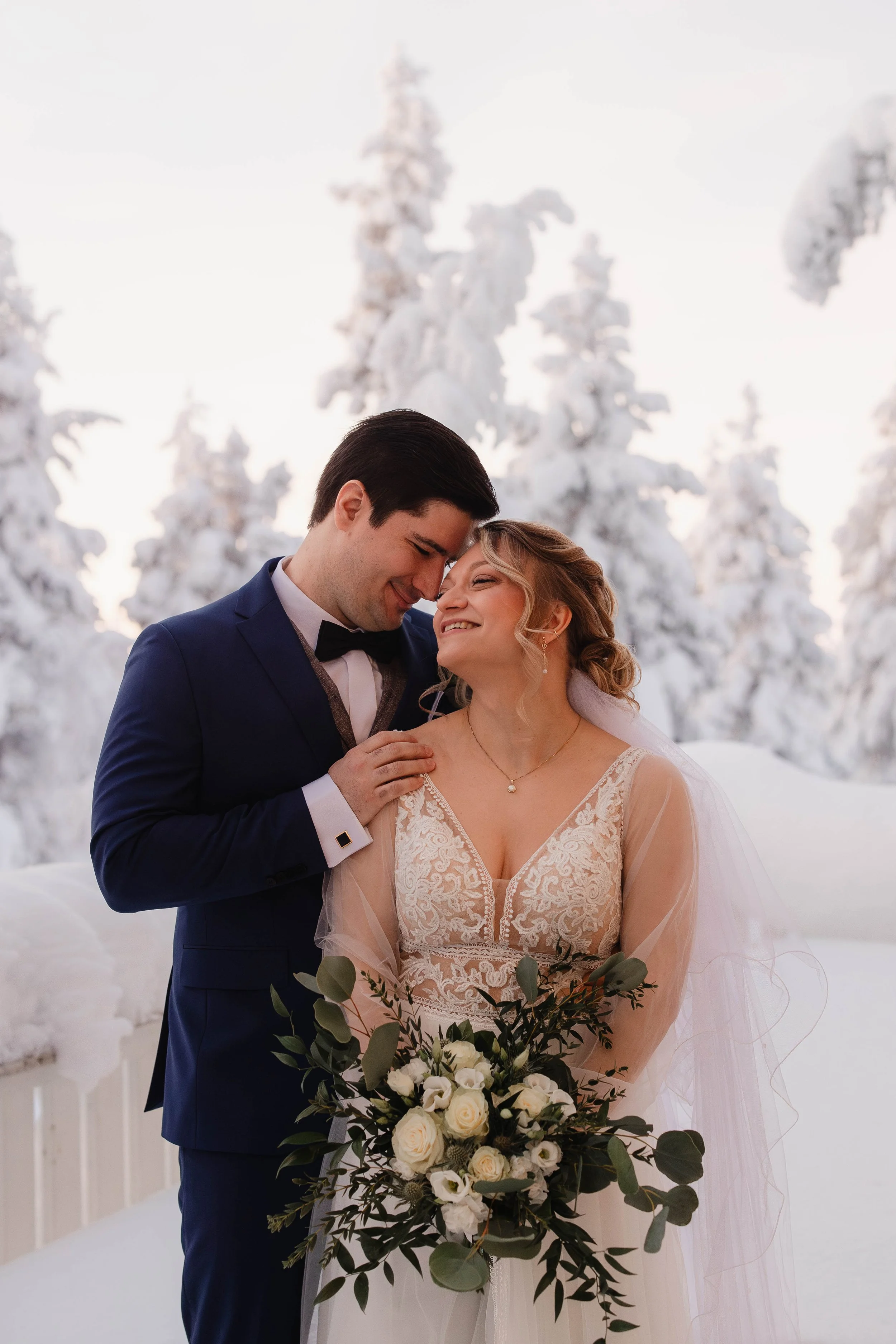 A newlywed couple in wedding attire smiling and leaning their foreheads together outdoors in a snowy winter landscape, with snow-covered trees in the background, and the bride holding a bouquet of white roses. Tina the Lapland Photographer.