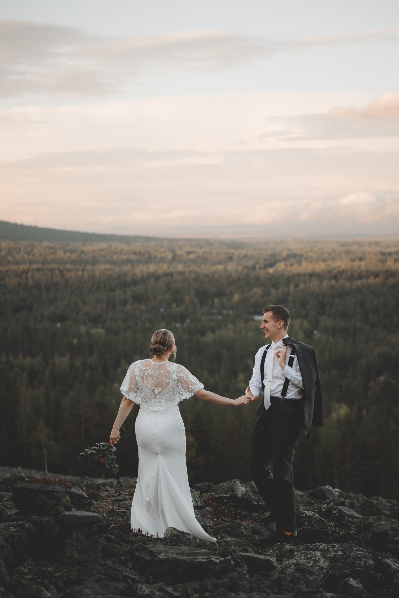 A bride and groom holding hands outdoors on rocky terrain with a scenic mountain view and sky in the background.