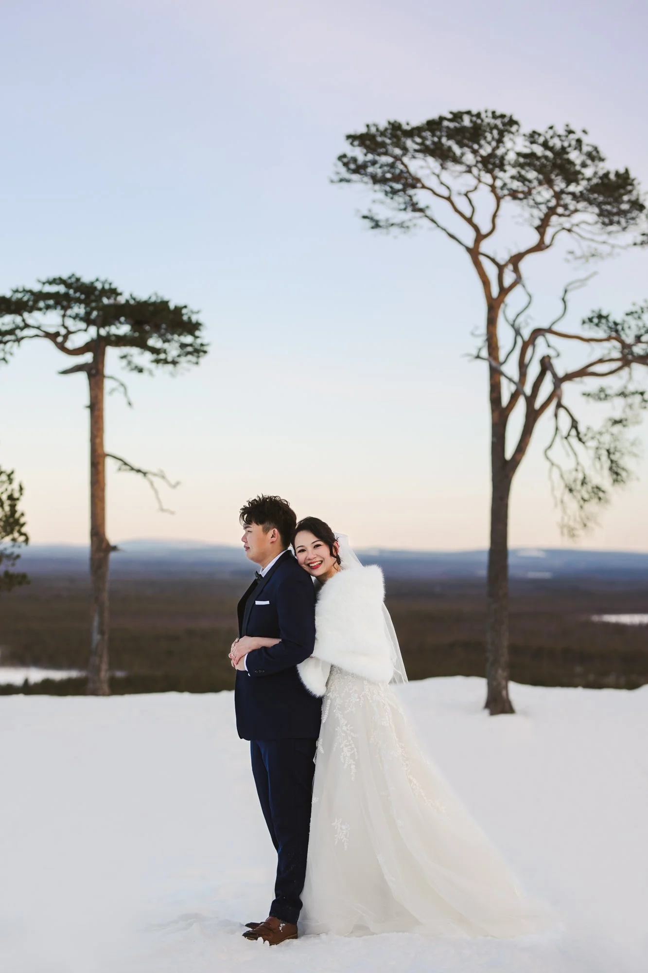 Wedding photos Lapland. A bride and groom standing closely together in a snowy landscape with tall, sparsely leaved trees in the background during sunset.