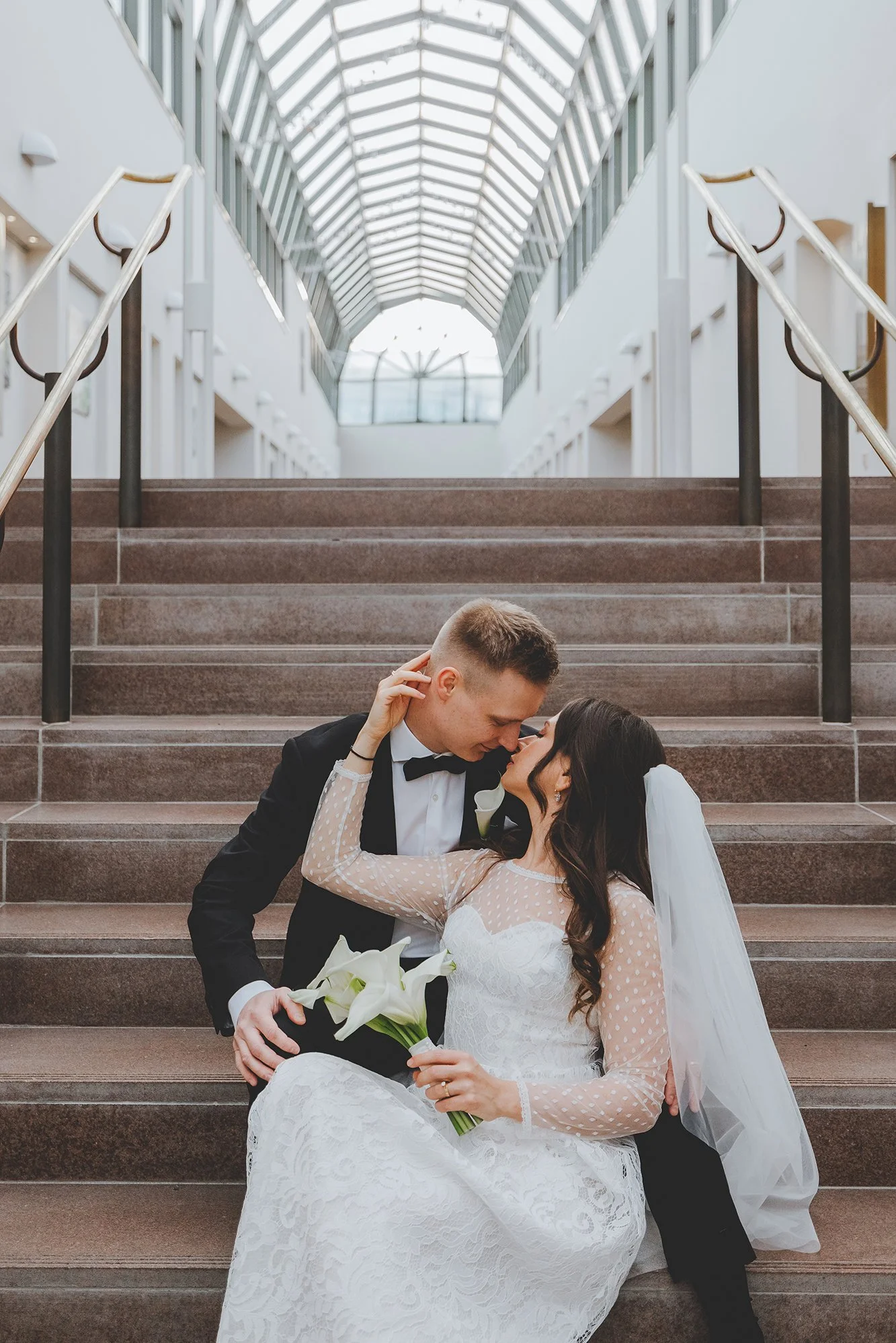 Wedding photographer Rovaniemi. A newlywed couple sits on the stairs inside a modern building with large windows and a skylight, sharing an intimate moment and smiling at each other.