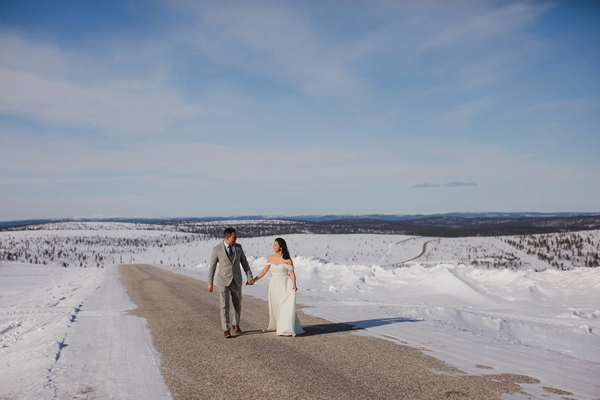 A couple dressed in wedding attire walking hand in hand on a snow-covered road in a winter landscape. Wedding photoshoot in Lapland.
