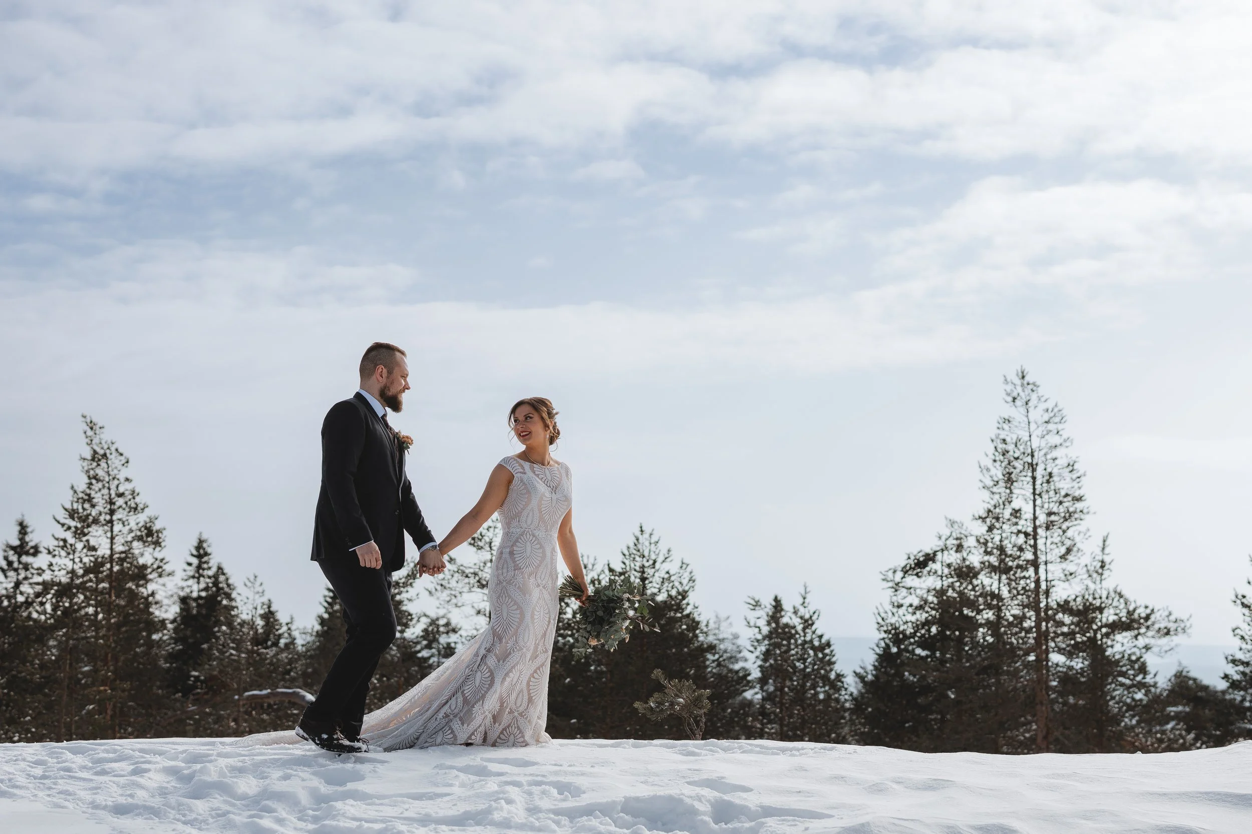 A bride and groom holding hands in a snowy outdoor setting, with the bride in a white lace wedding gown and the groom in a black suit, surrounded by tall trees and a cloudy sky. Tina the Lapland Photographer.