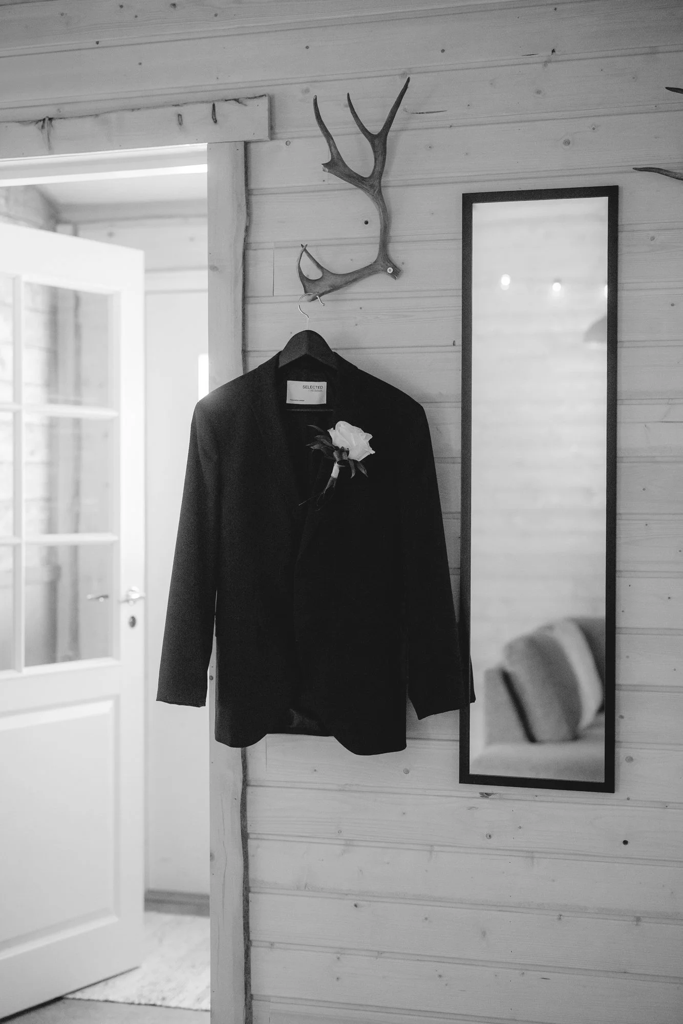 Black suit jacket with a white flower pinned on it hanging on a hanger, reflected in a full-length mirror on a wooden wall.