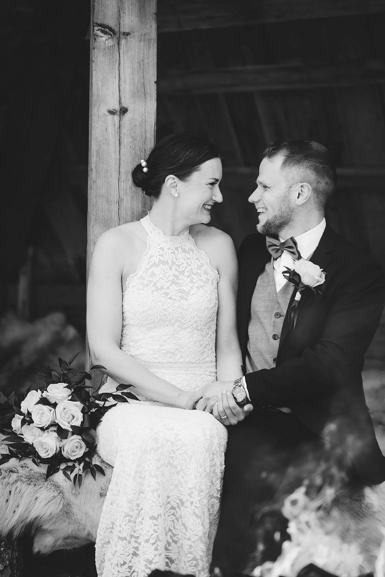 A black-and-white photo of a couple at their wedding, holding hands and smiling at each other. The bride wears a lace dress and has dark hair styled in an updo, and the groom wears a dark suit with a bow tie and a boutonniere. There is a large bouque