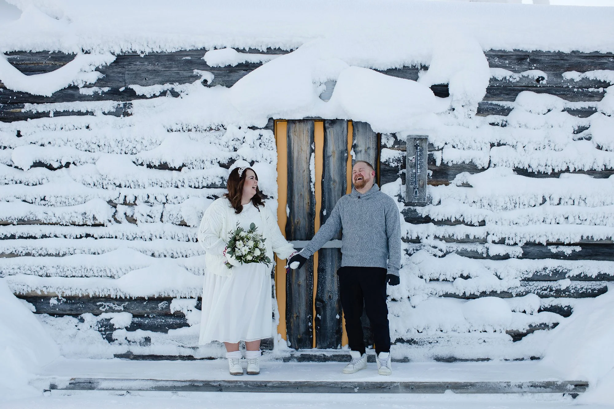 A bride and groom holding hands, standing in front of a snow-covered wooden wall, smiling and laughing during their winter wedding in Rovaniemi.