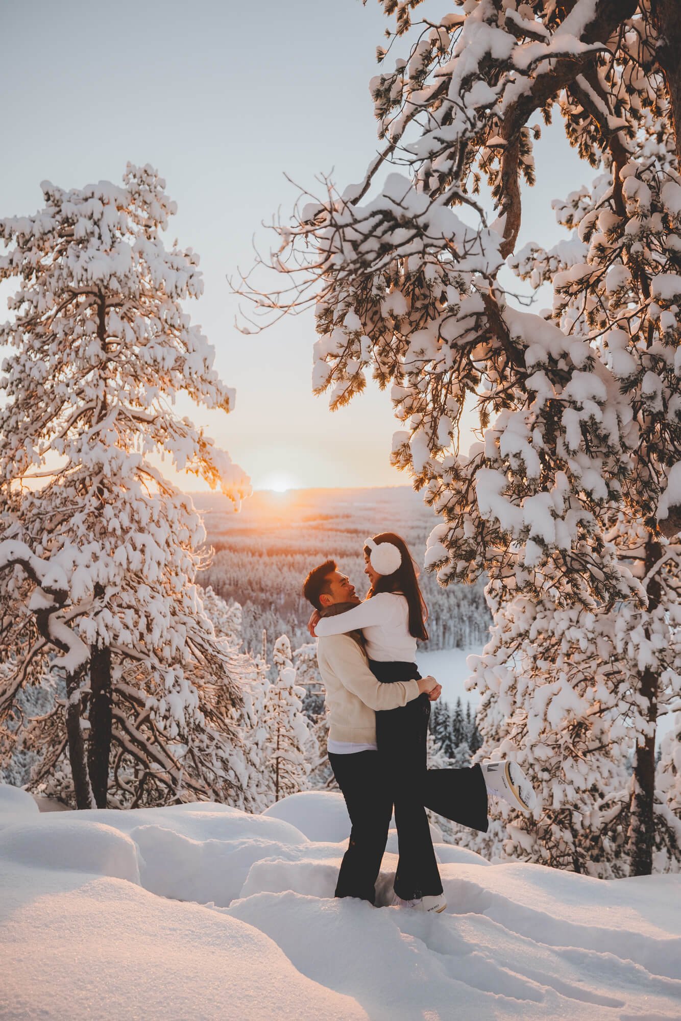 A couple is standing in a snowy landscape, with the man lifting the woman off the ground as they face each other and smile. They are surrounded by snow-covered trees, and the sun is setting in the background, casting a warm glow over the scene.
