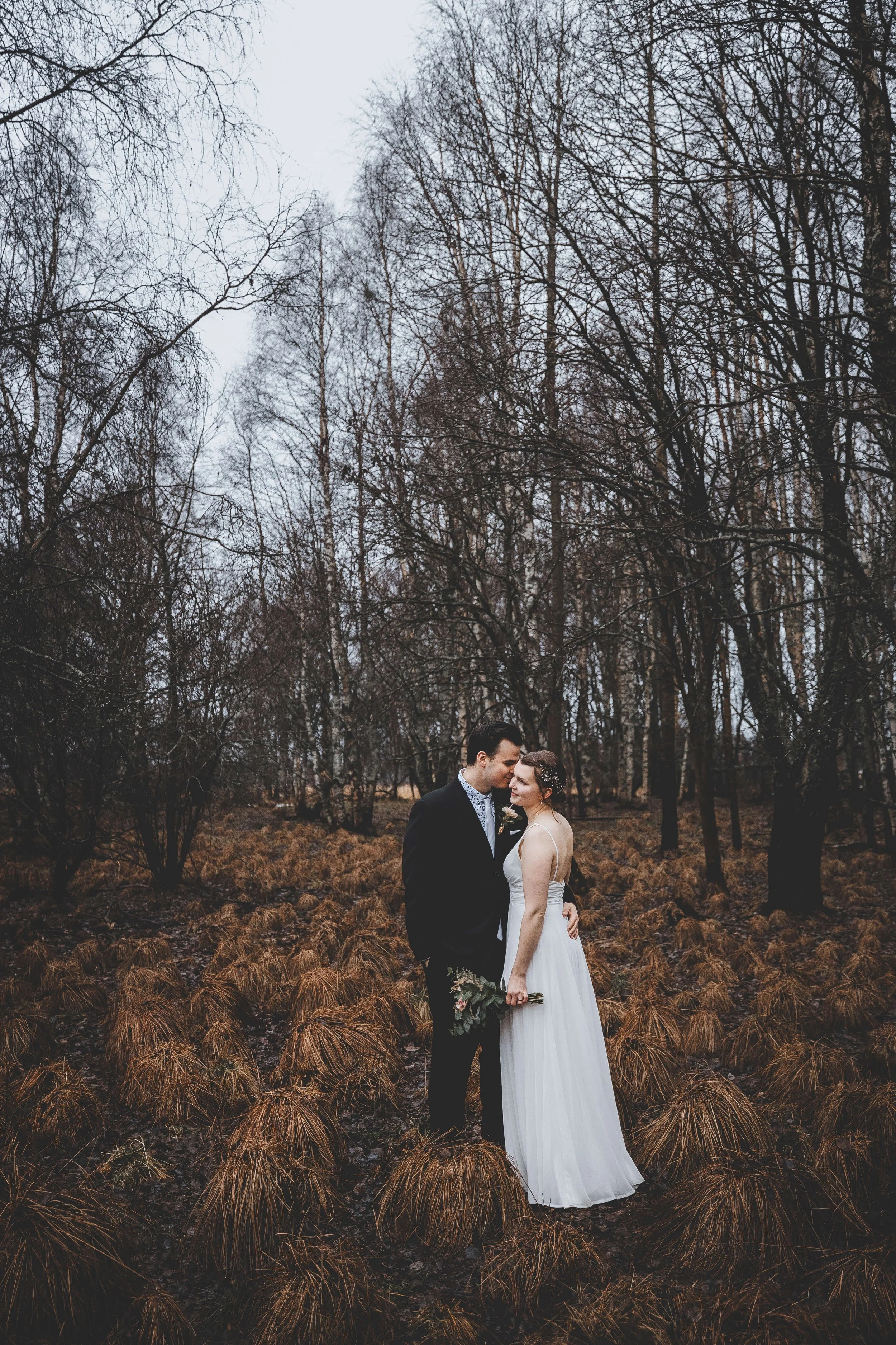 A bride and groom standing close together in a wooded area, holding hands and looking into each other's eyes. The bride is in a white gown holding a bouquet, and the groom is in a black suit. Bare trees and brown ground cover surround them.